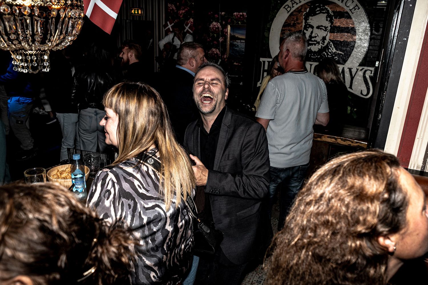 Laughing man in a dark jacket stands among a lively crowd of people socializing at a crowded, dimly lit pub with a flag and wall poster.