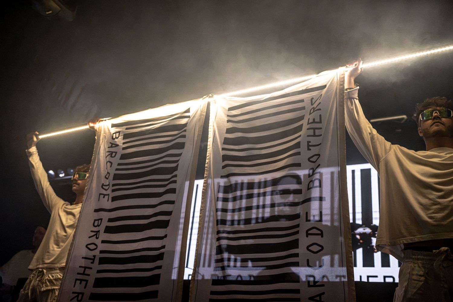 Performers hold up Barcode Brothers banner on foggy concert stage with bright LED lights.