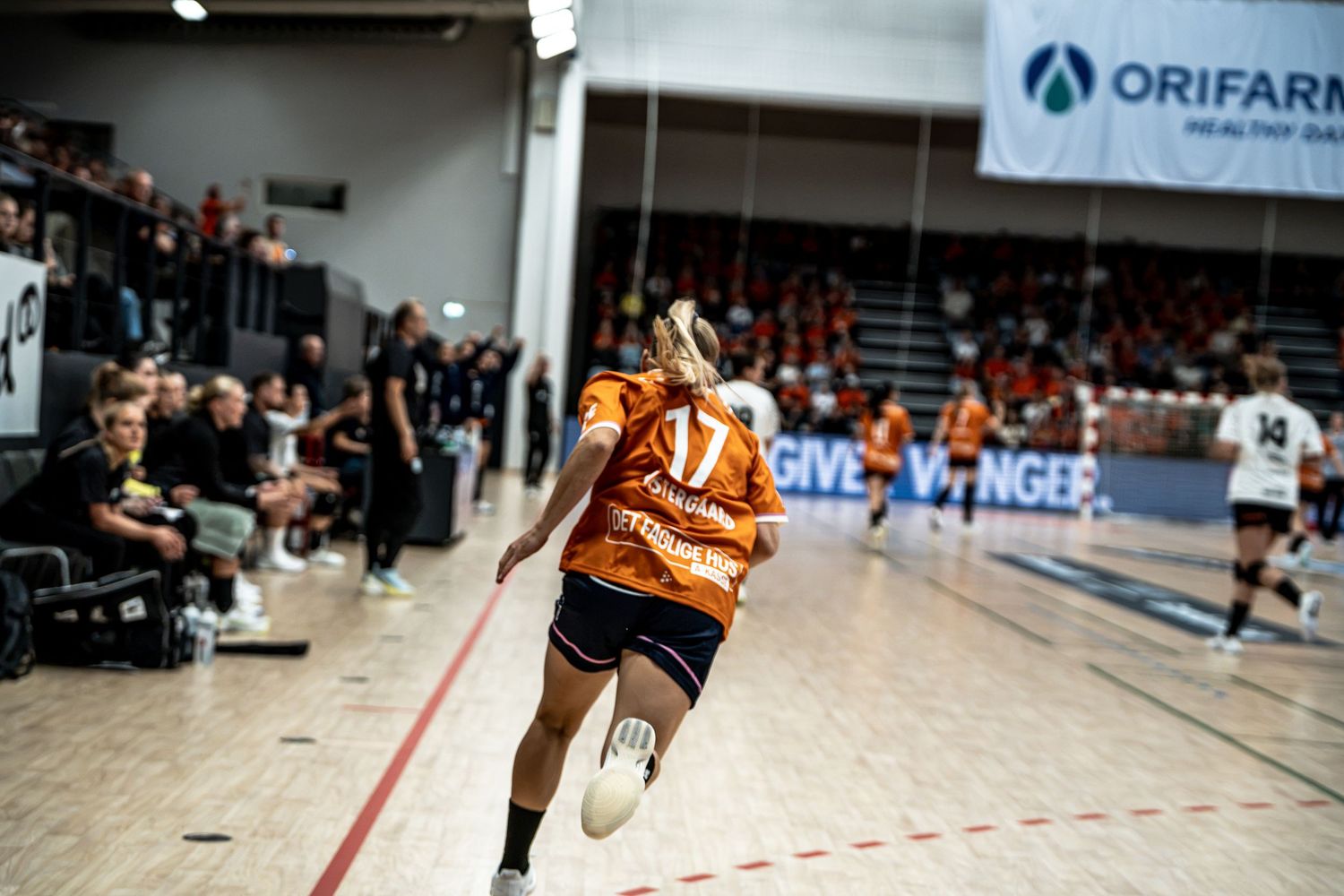 Female handball player in orange jersey runs along the sideline during an indoor handball match.