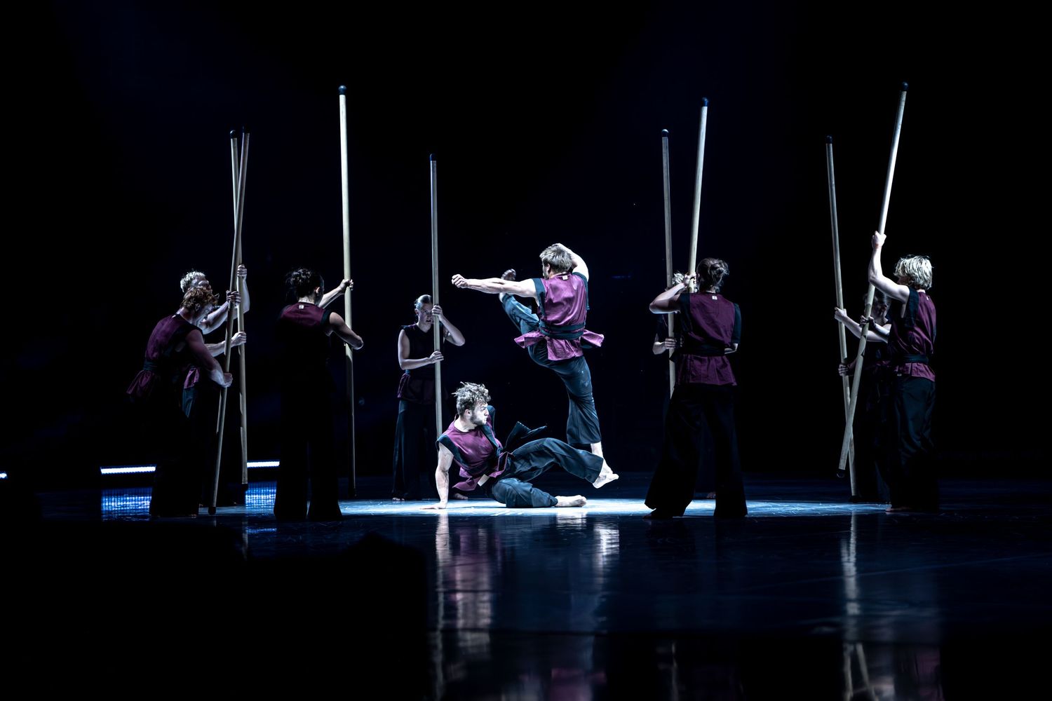 Group of dancers in burgundy costumes perform choreographed routine with tall poles on stage.