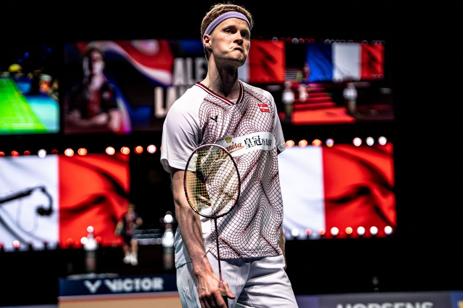 Danish badminton player Anders Antonsen standing on court with French flag displayed on arena screen behind him.