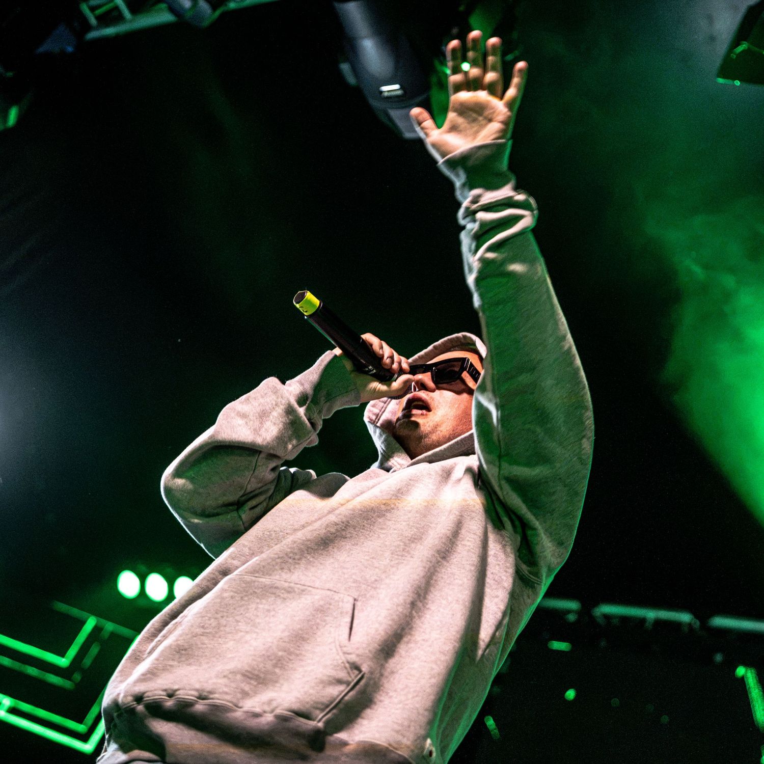 Rapper performing on stage in grey hoodie with hand raised under green stage lights.