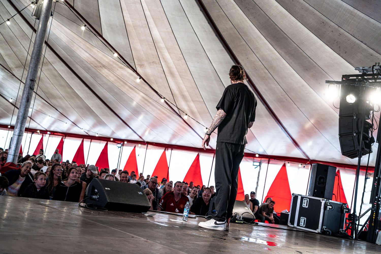 Tattooed performer stands before large crowd at outdoor music festival under striped tent canopy.