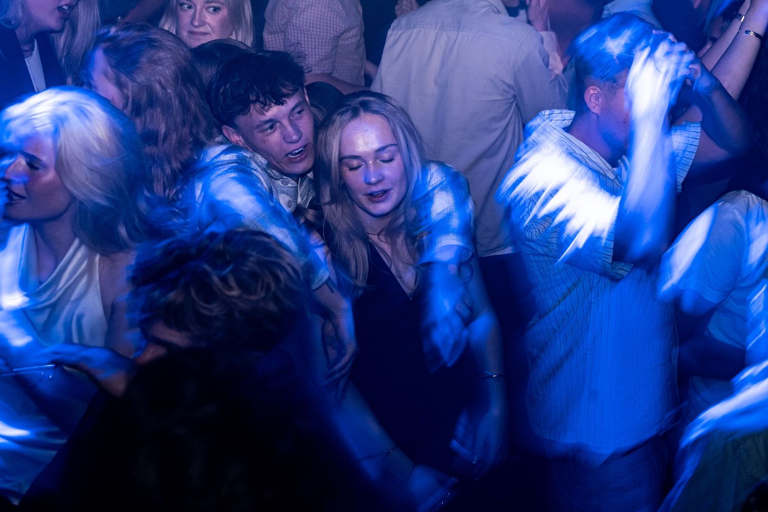 Partygoers dance close together in a crowded nightclub under blue and purple lights, a young woman and man in the center.
