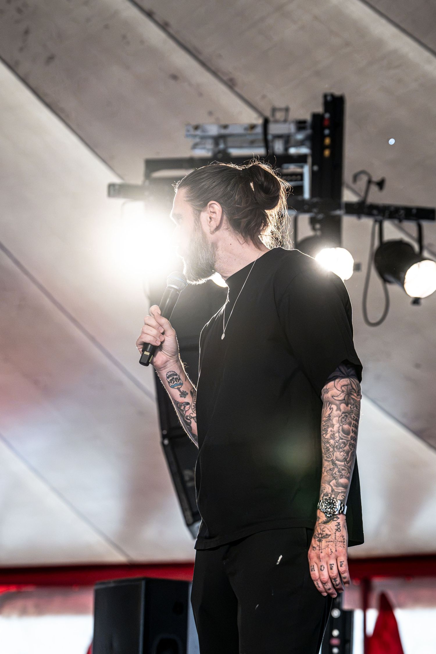 Heavily tattooed bearded male performer holds microphone under bright stage lights at music festival.
