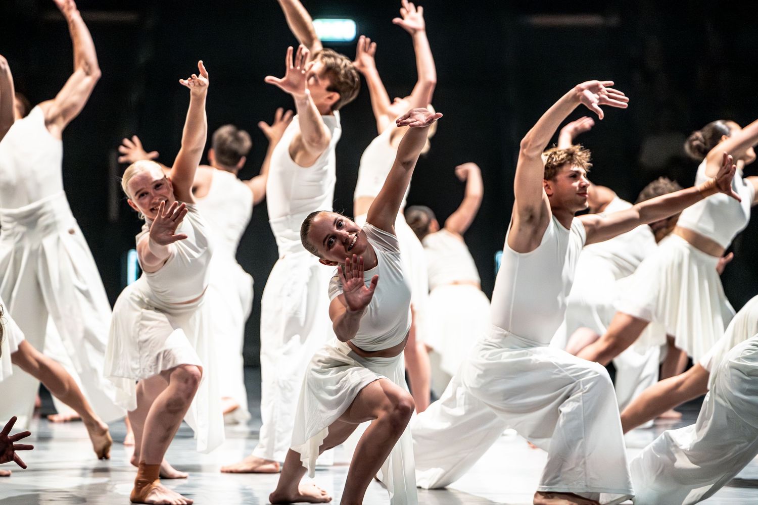 Large group of dancers in white costumes performing dynamic choreography on brightly lit stage.