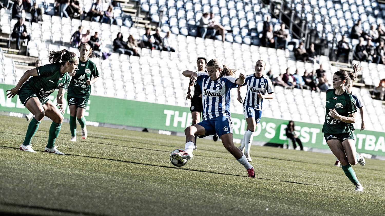 Women's soccer match action as blue-white player dribbles past green-uniformed defenders on stadium pitch.