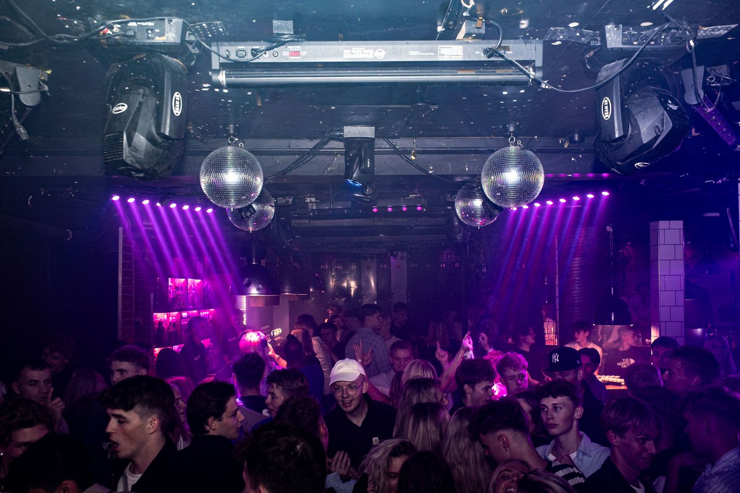 Packed nightclub dance floor under shimmering disco balls and glowing purple lights as the crowd enjoys the party.