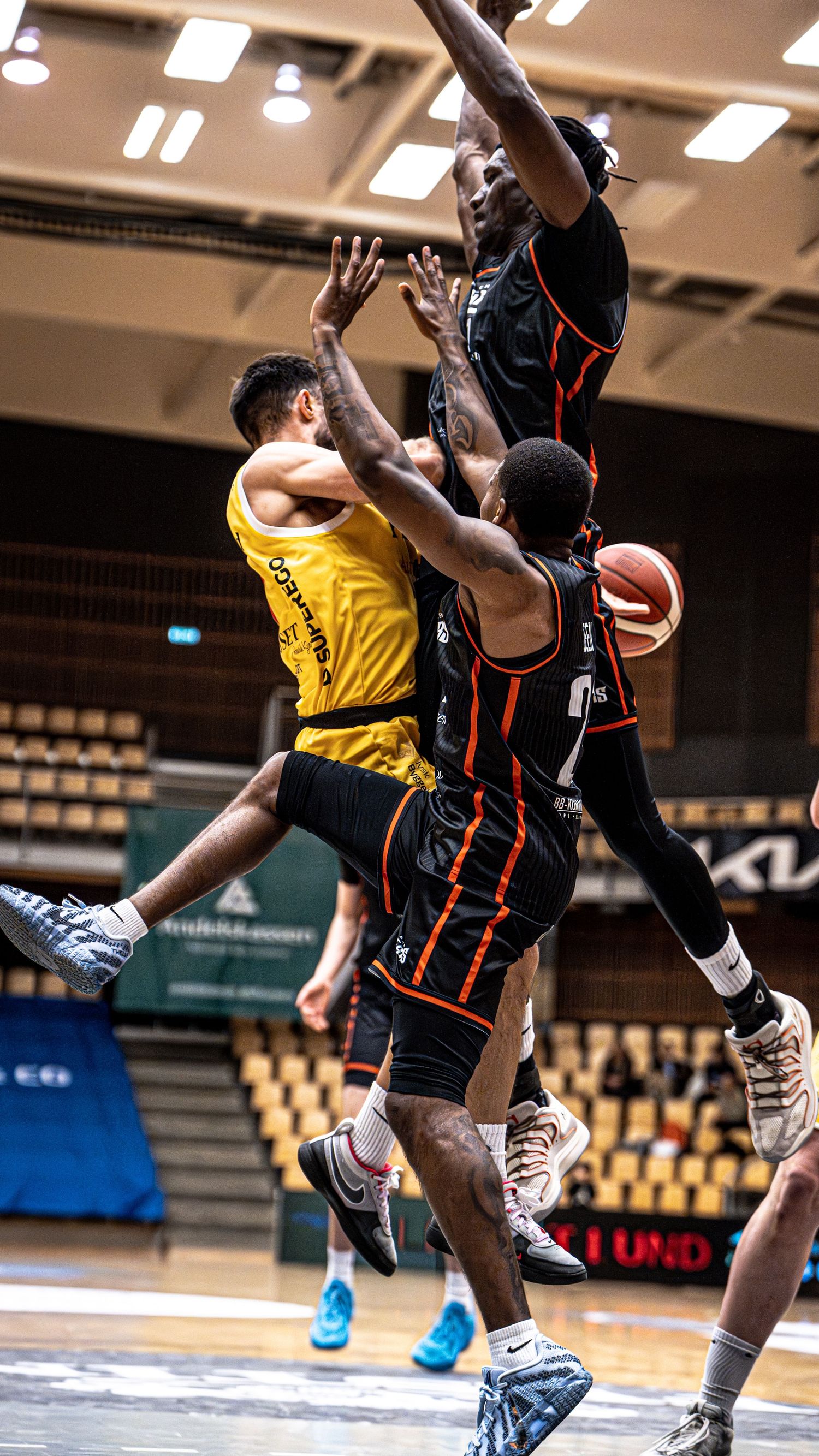 Basketball player in yellow jersey drives between two defenders in black uniforms.
