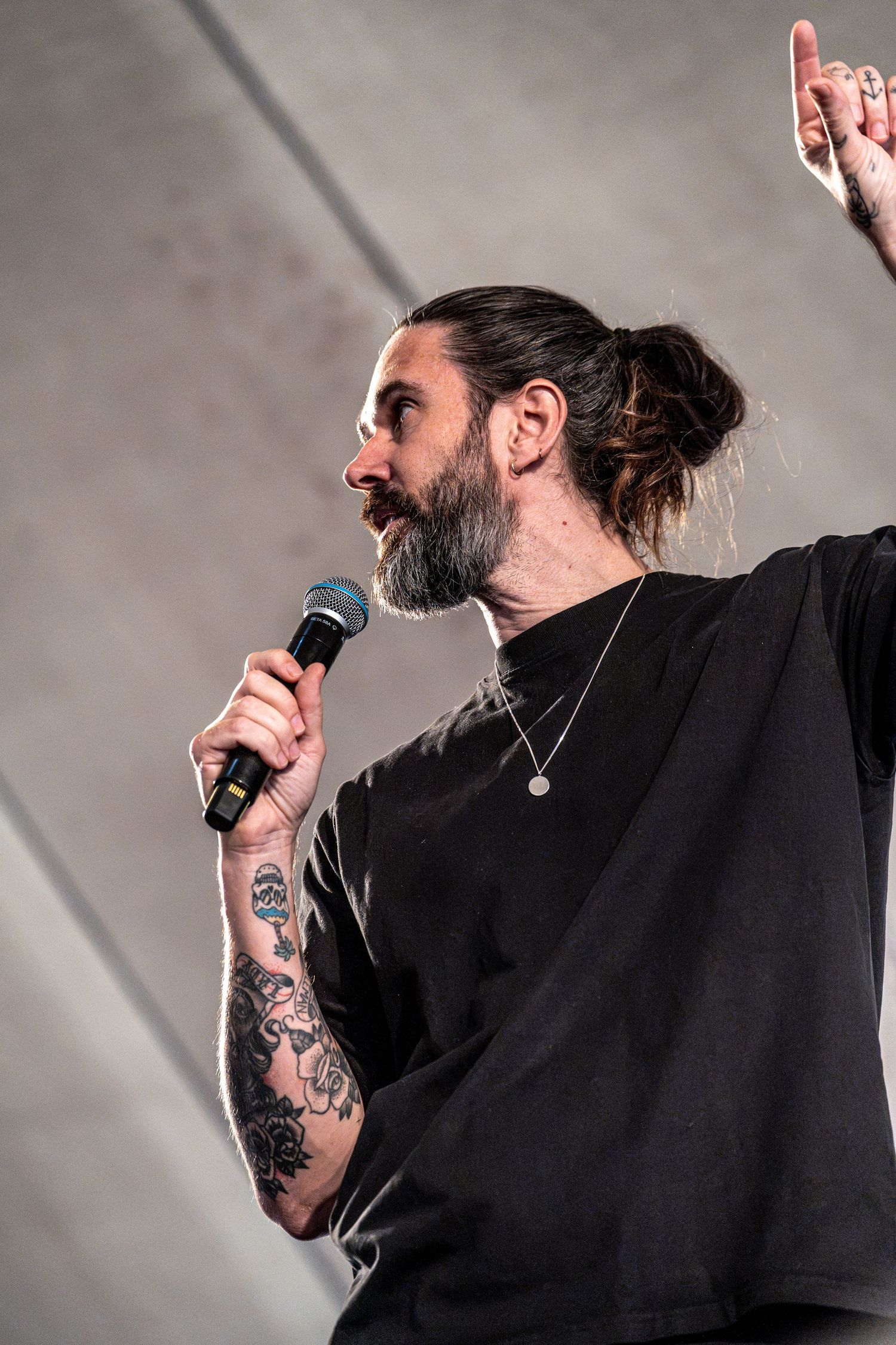 Side profile of tattooed bearded singer with hair bun performing on stage with microphone.