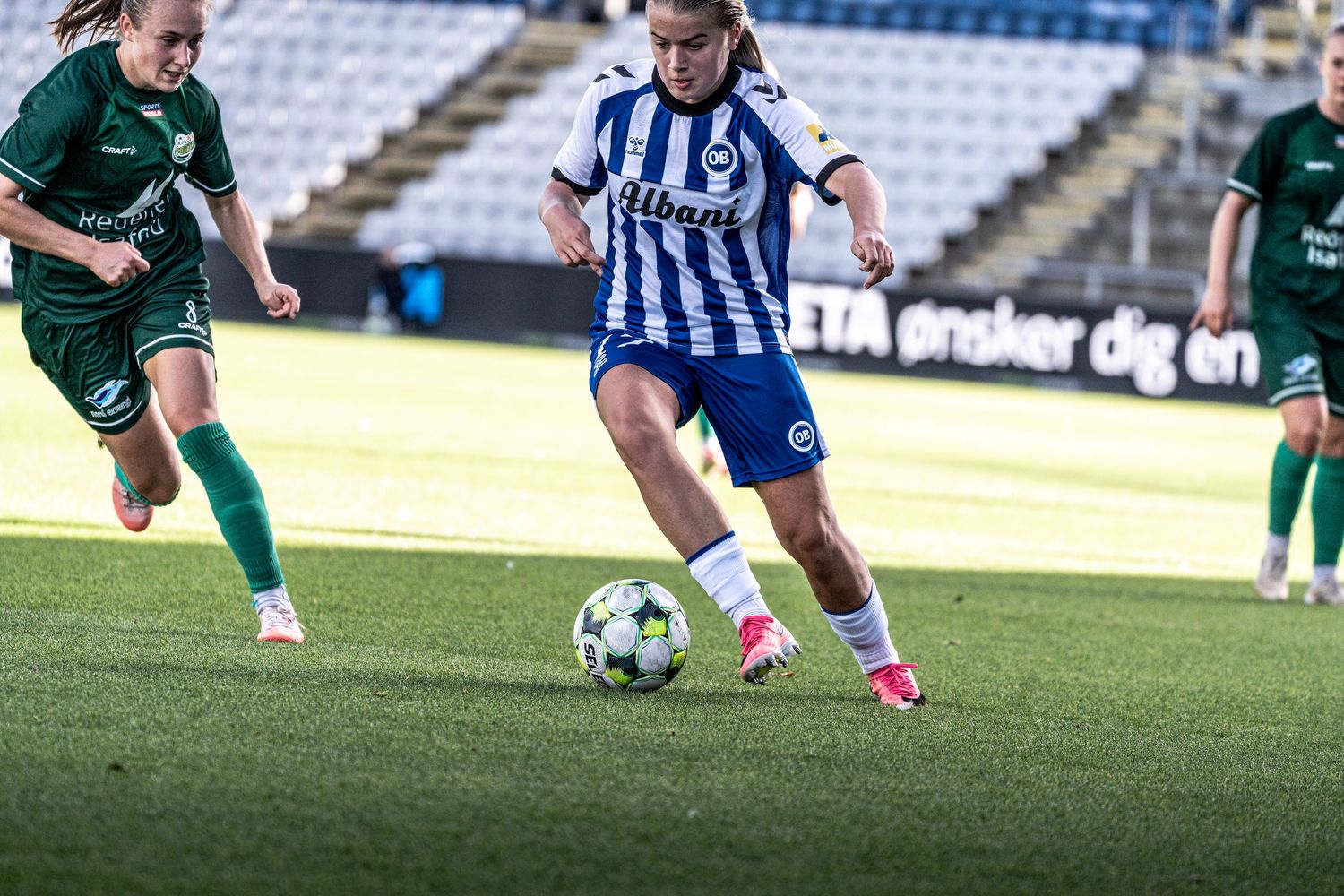 Women's soccer player in blue and white kit controls the ball while being pursued by a green-shirted opponent.