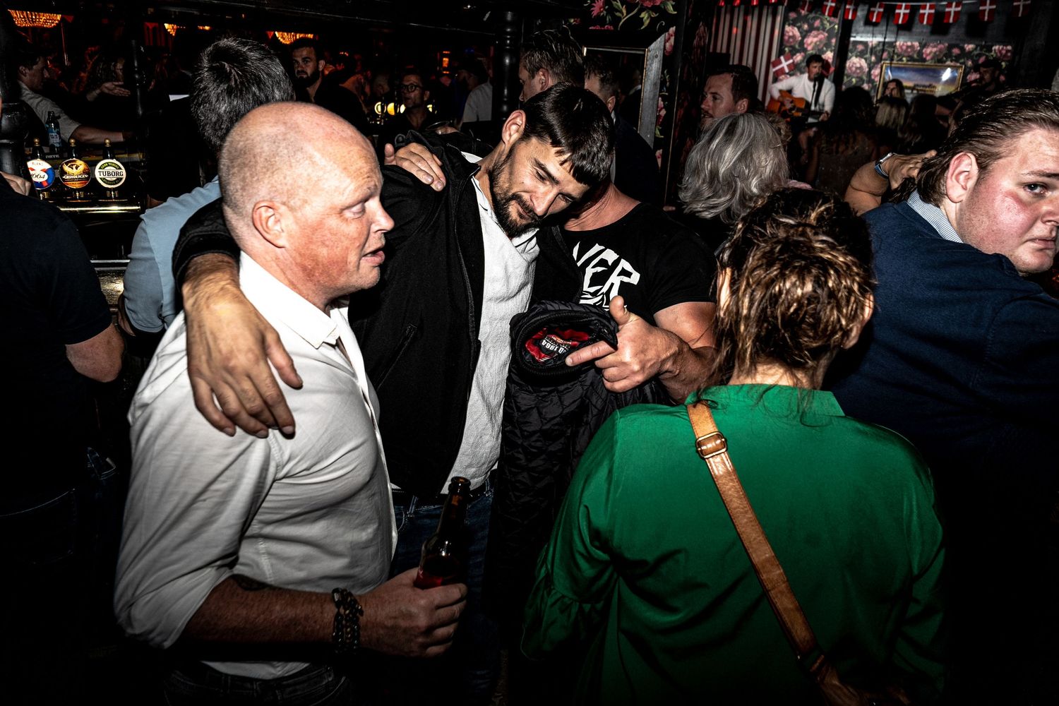 Bald man in a white shirt holds a drink while friends laugh and embrace in a crowded, dimly lit bar with a woman in green nearby.