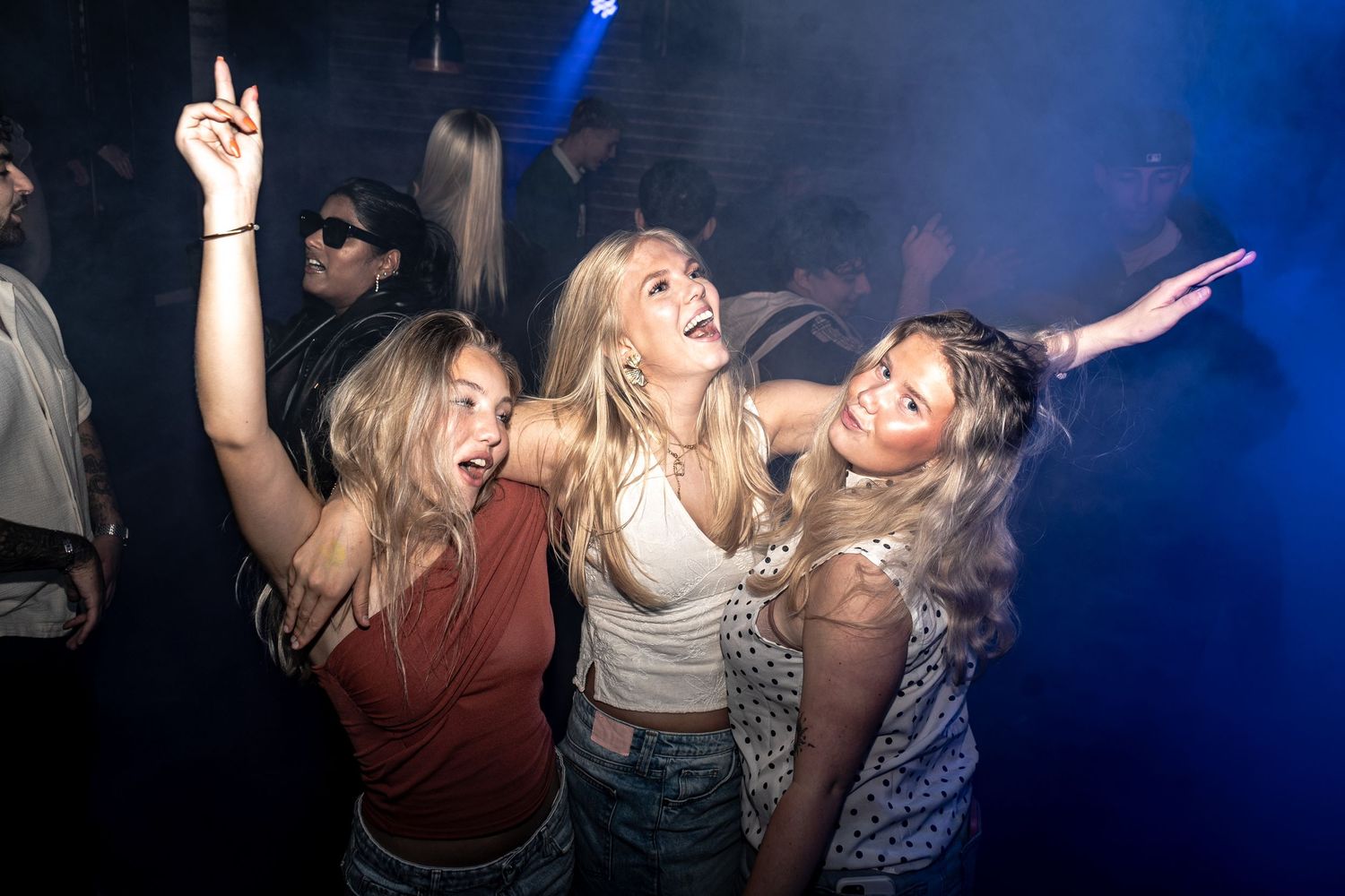 Three young women with long blonde hair dance and laugh together with raised arms on a crowded nightclub dance floor under blue lights.