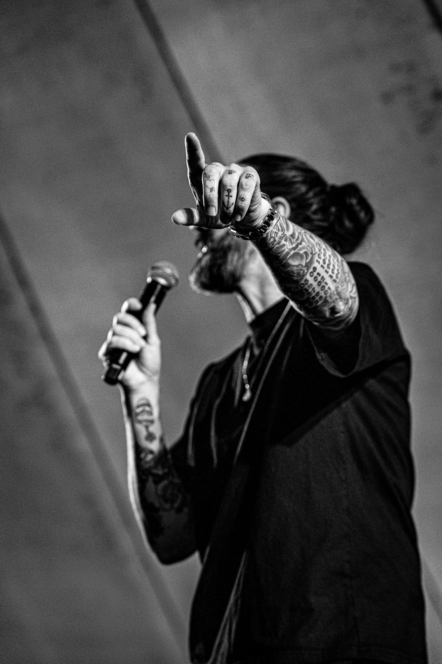 Tattooed performer in black pointing finger up while holding microphone on stage, black and white photo.