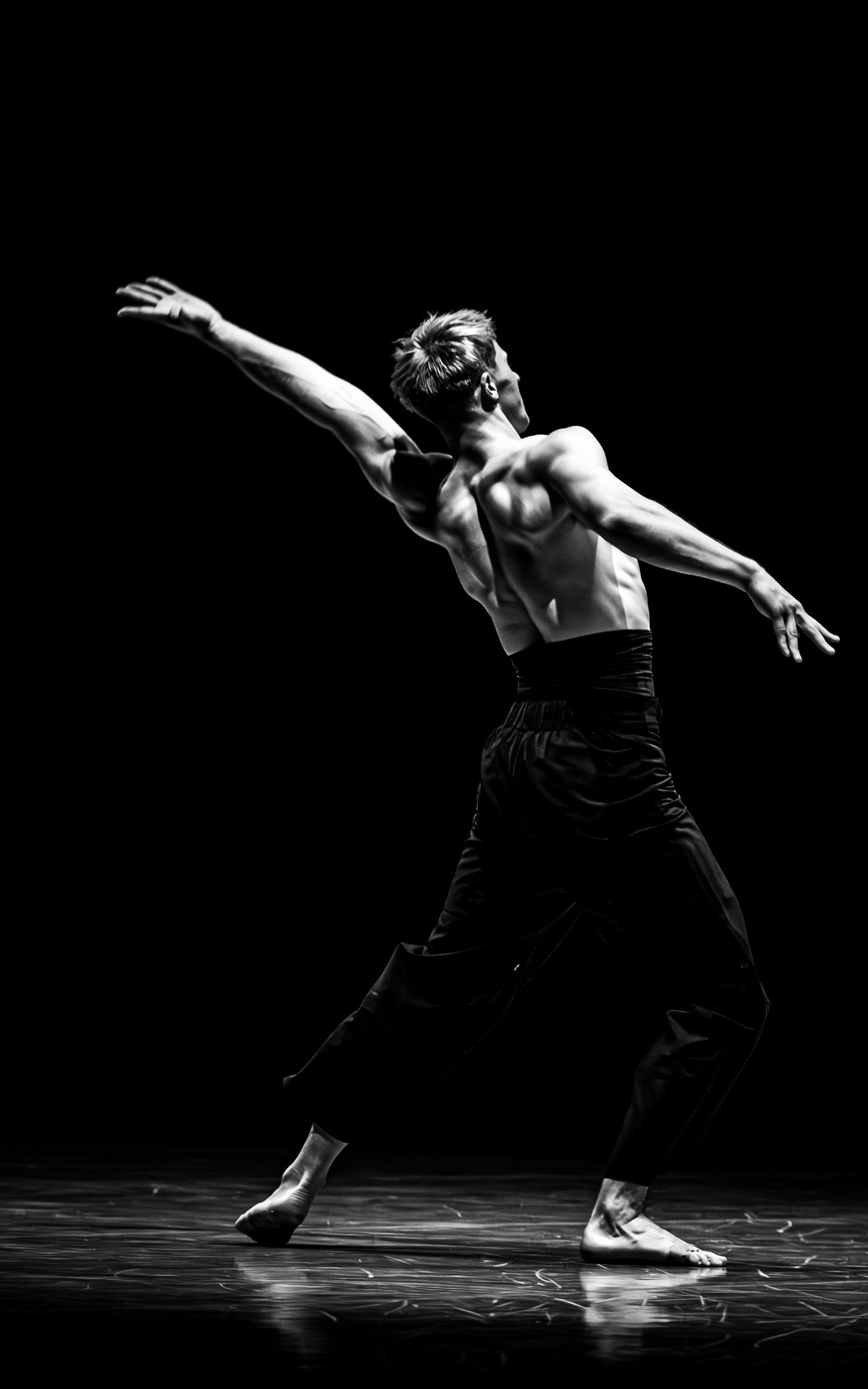 Shirtless male ballet dancer striking a dramatic pose on a dark stage in black and white.