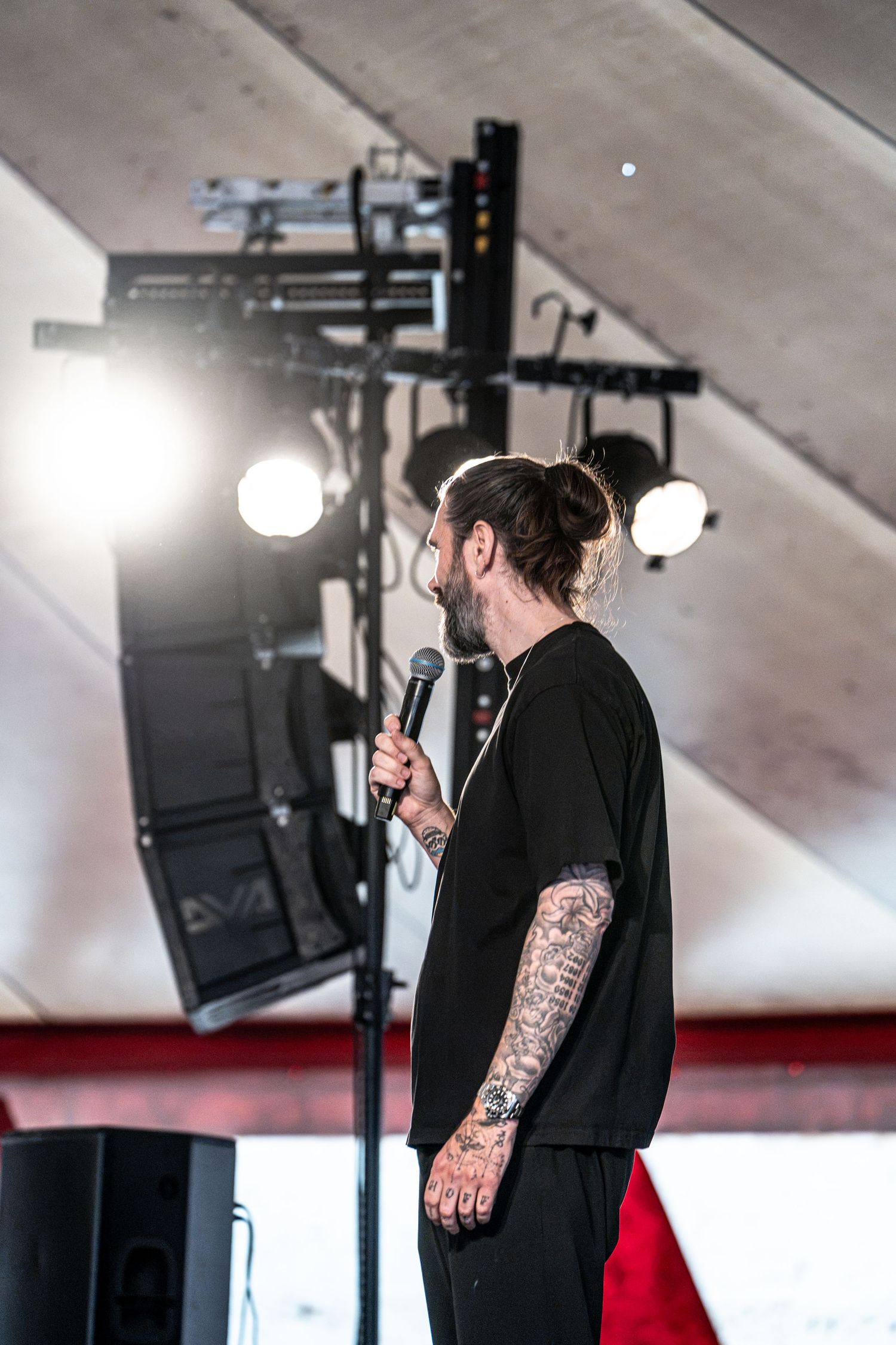 Side profile of tattooed male performer holding microphone stand on stage with stage lights.