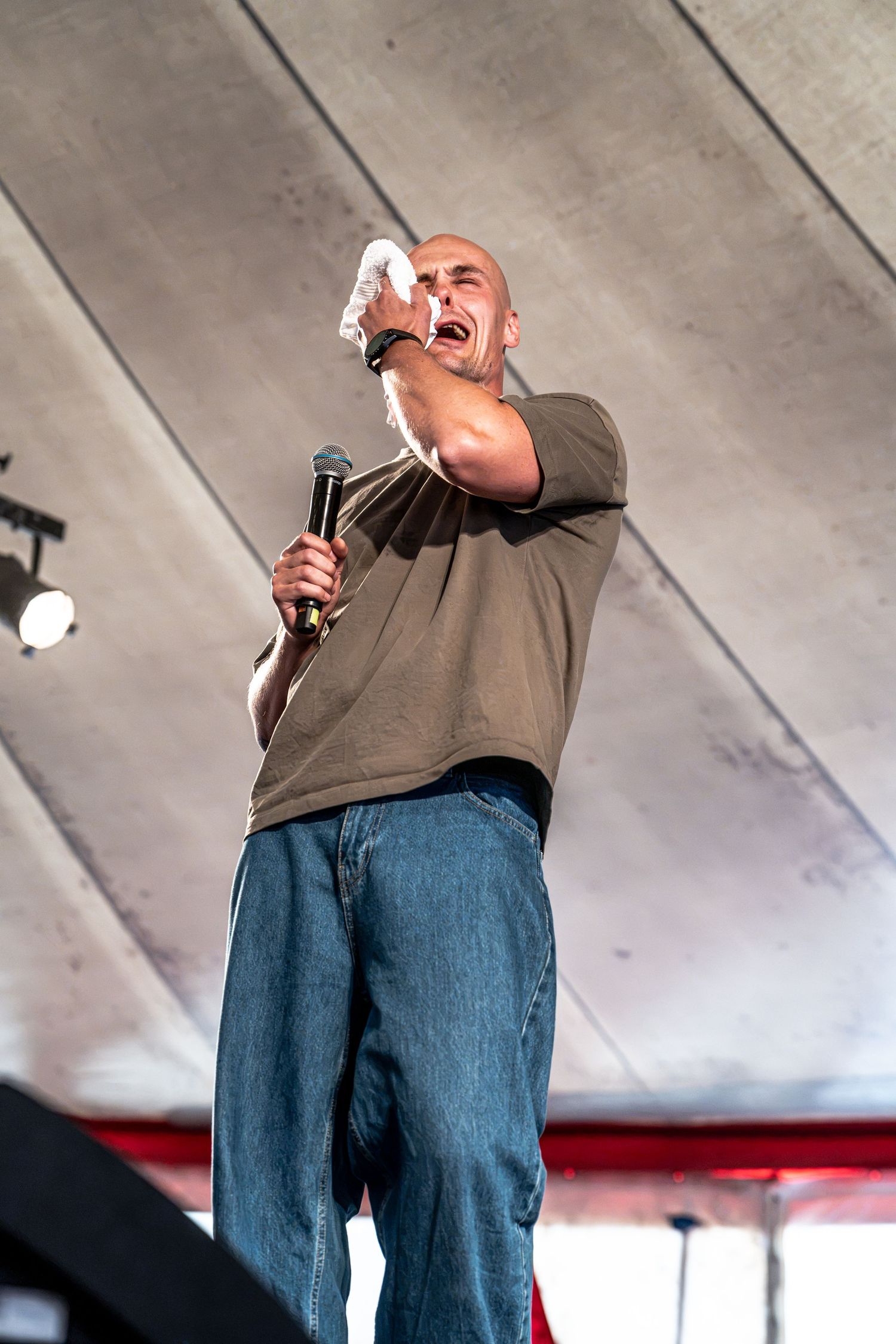 Bald male performer points at crowd while shouting into microphone on stage at outdoor festival event.