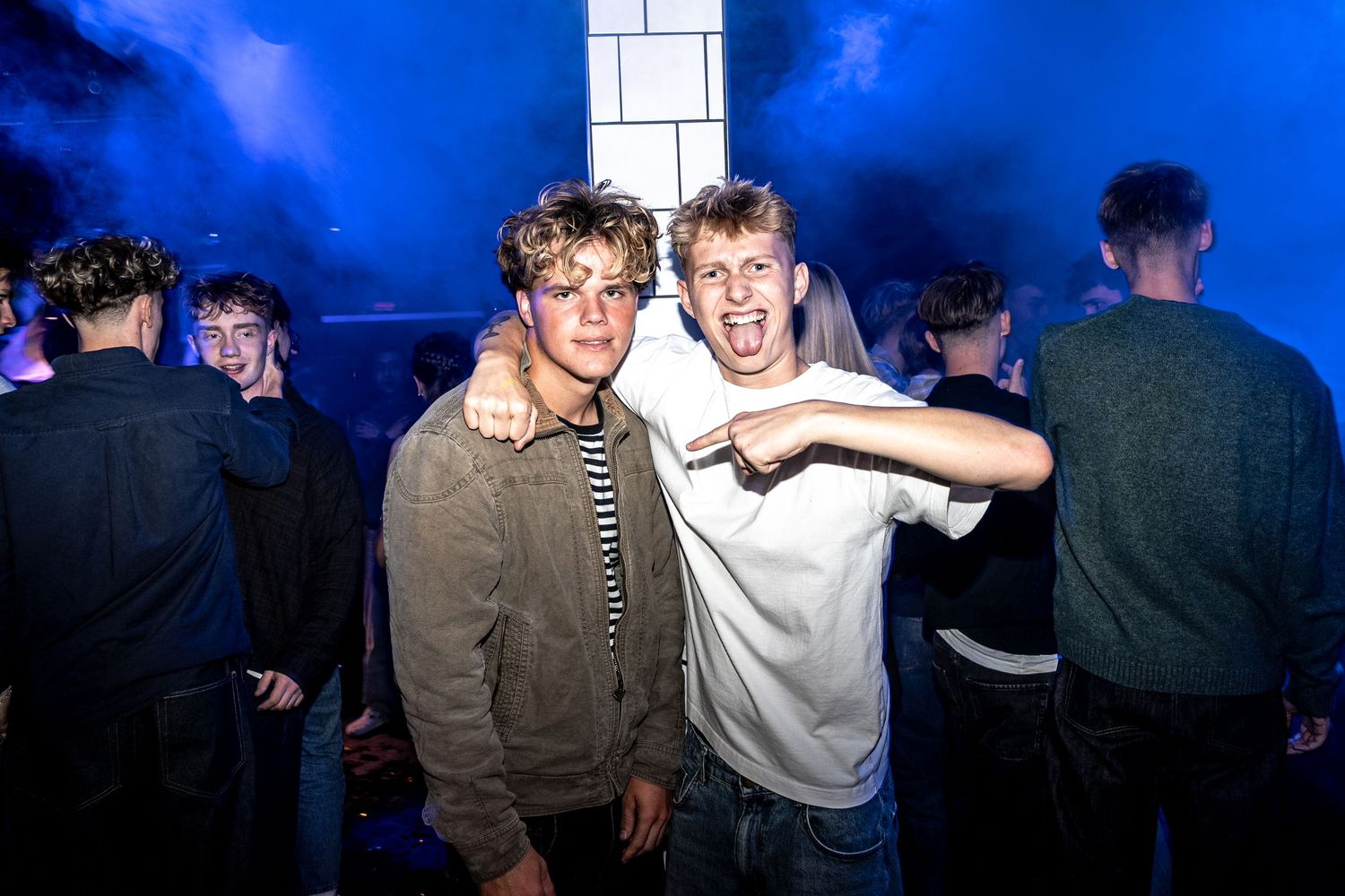Two young men pose together with arms around each other at a nightclub party, surrounded by a crowd under blue stage haze.
