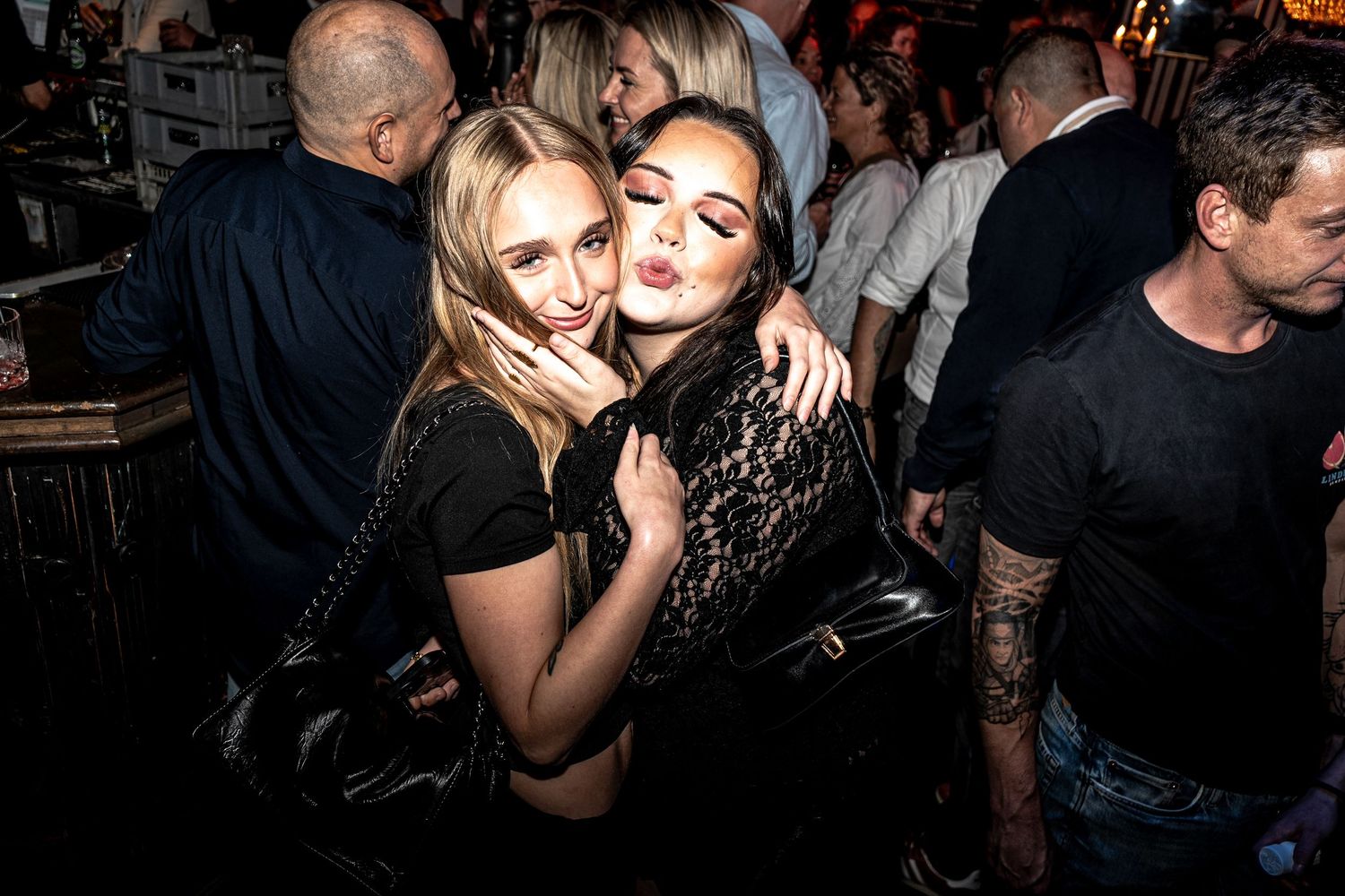 Two smiling young women embrace and pose cheek to cheek at a crowded nightclub while other guests socialize around them.