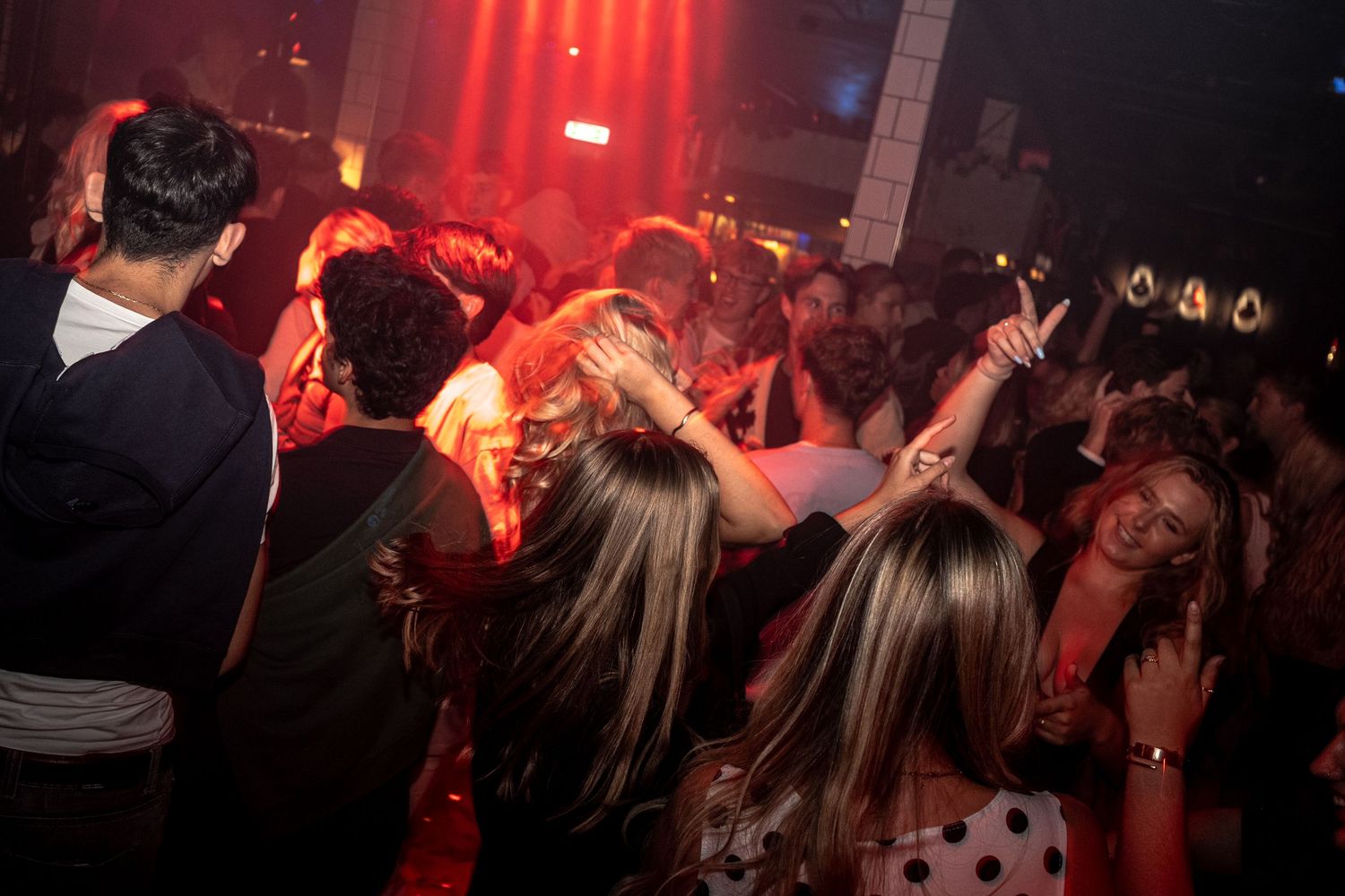 Crowd of partygoers dancing with raised arms in a dimly lit nightclub with glowing red lights overhead.