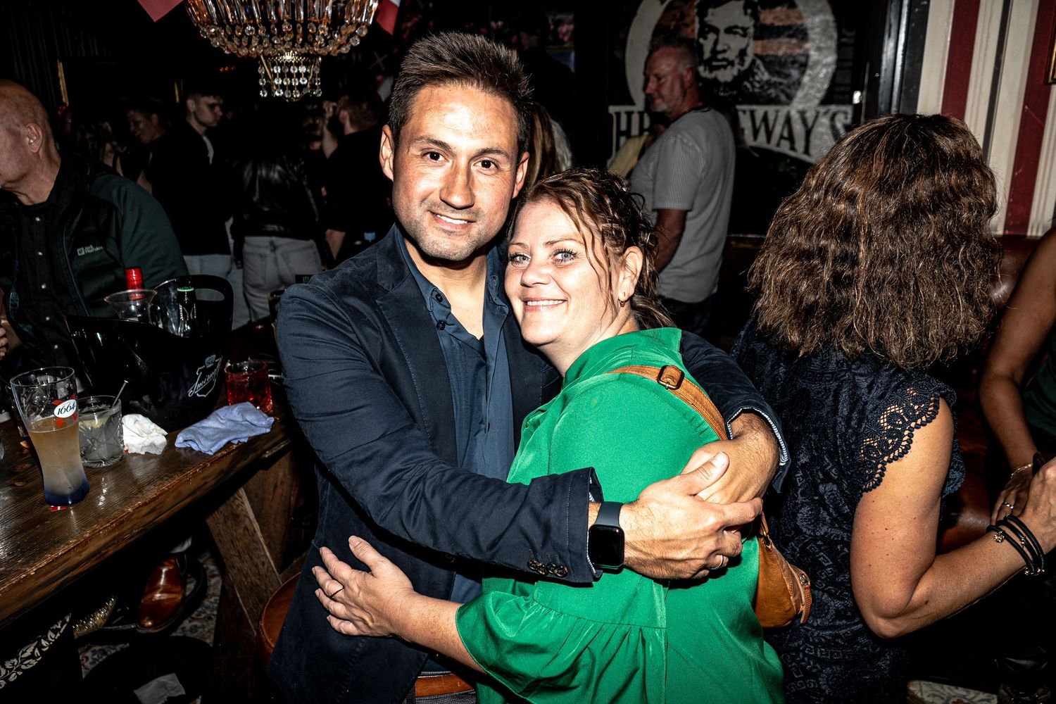 A smiling man in a dark blazer hugs a woman in a green dress at a lively bar, with guests chatting in the background.