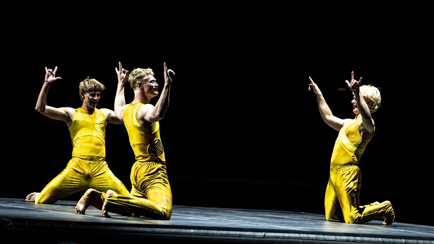 Three performers in bright yellow costumes kneel and gesture on stage during dance performance.