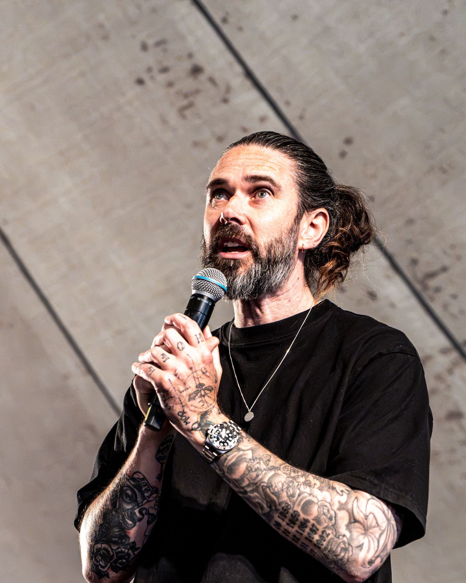 Heavily tattooed bearded man with hair bun speaking into microphone wearing black t-shirt outdoors.
