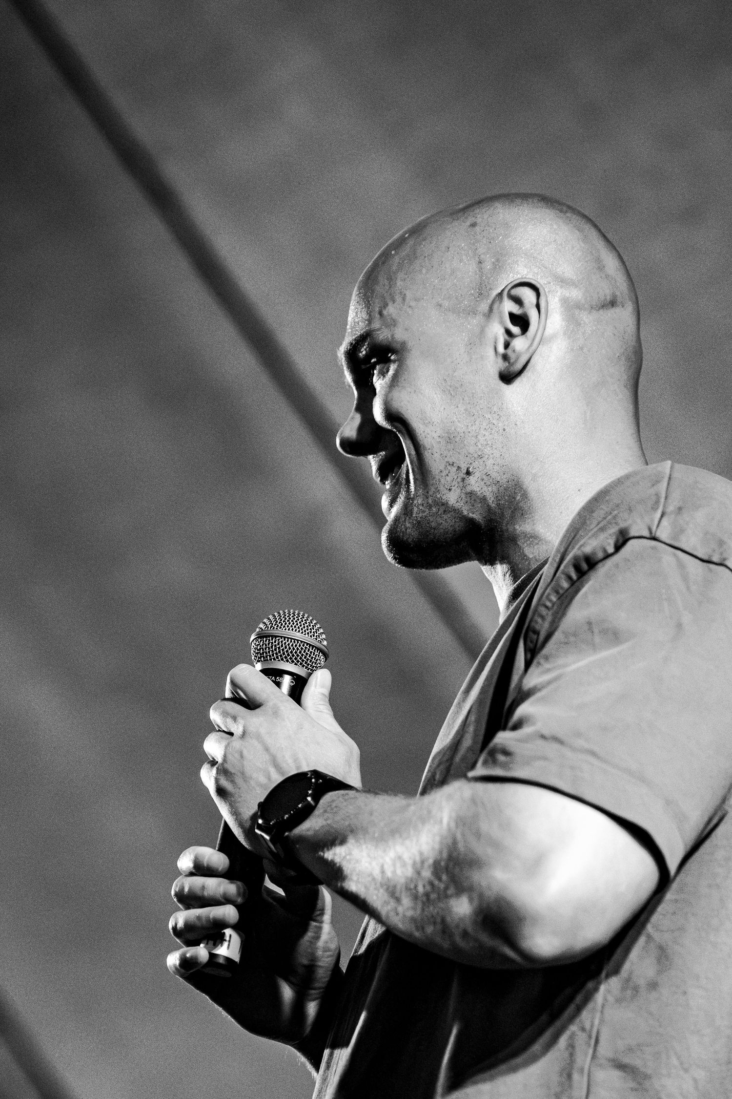Black and white profile shot of a bald male singer performing passionately into a microphone on stage.