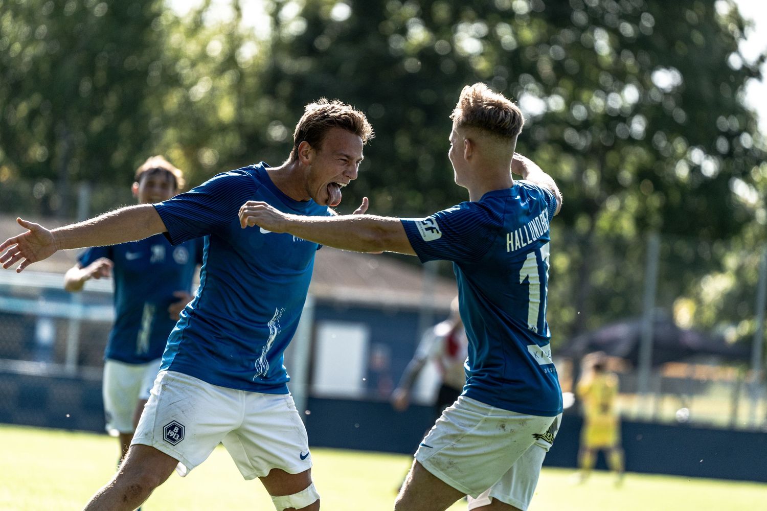 Two soccer players in blue jerseys celebrate a goal together during a sunny match.