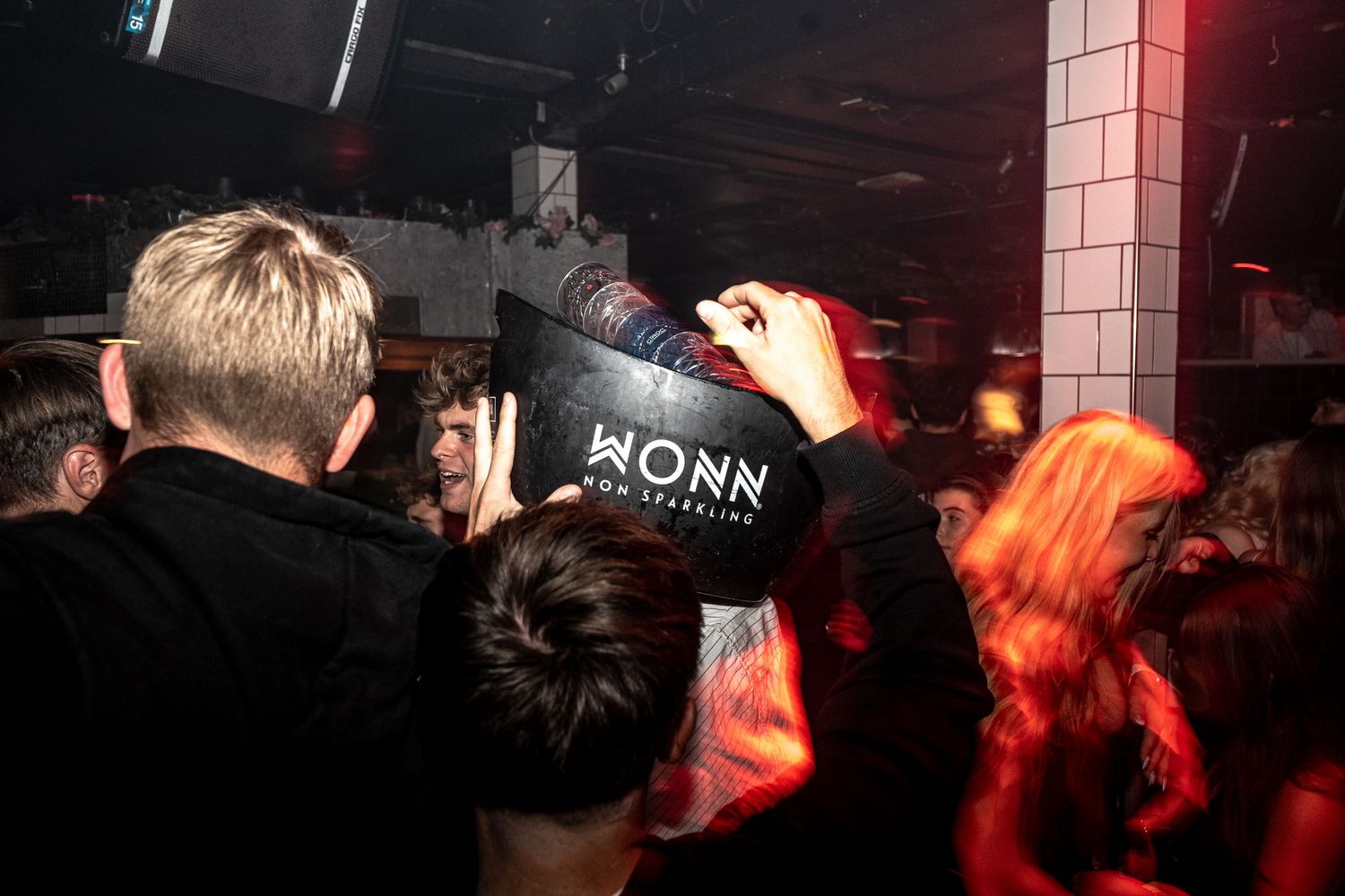 Partygoers in a dark club watch a person holding a Wonn sparkling sign, with red lighting and a brick pillar behind.