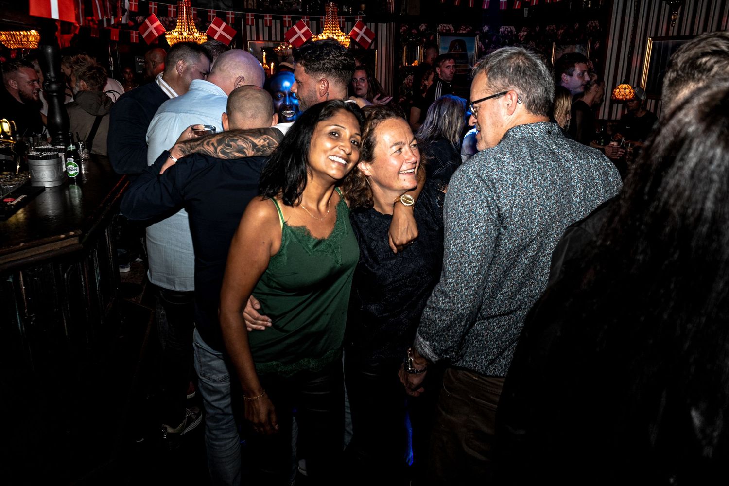Two smiling women pose close together in a busy, warmly lit bar while other guests chat around them.