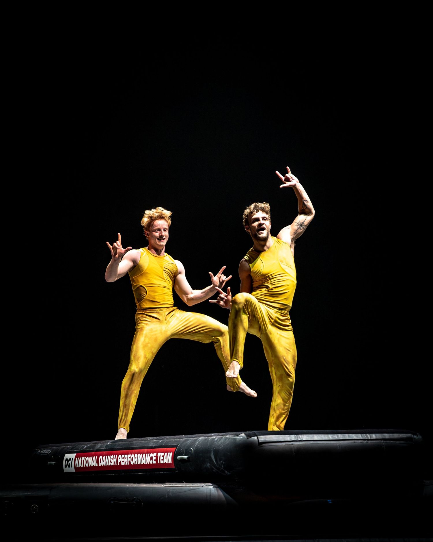 Two male gymnasts in yellow suits celebrate on National Danish Performance Team trampoline.