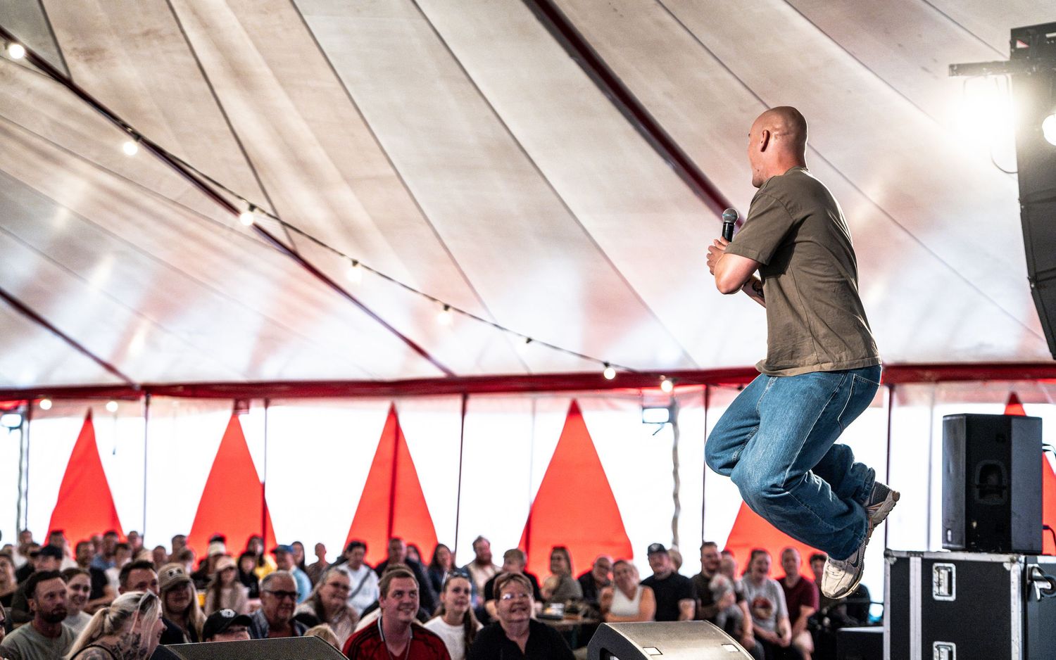Energetic performer jumps on stage before a large crowd under a white and red tent at an outdoor event.