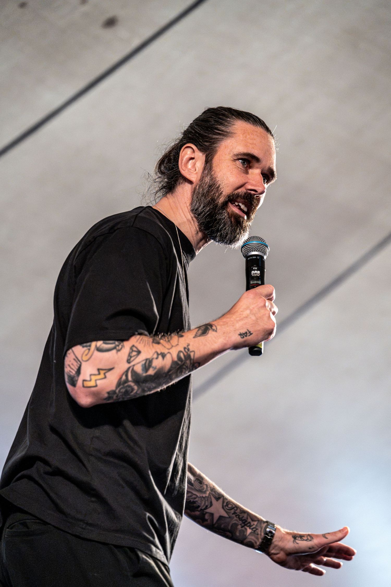 Bearded male performer with long hair and sleeve tattoos singing into microphone in a black t-shirt on stage.