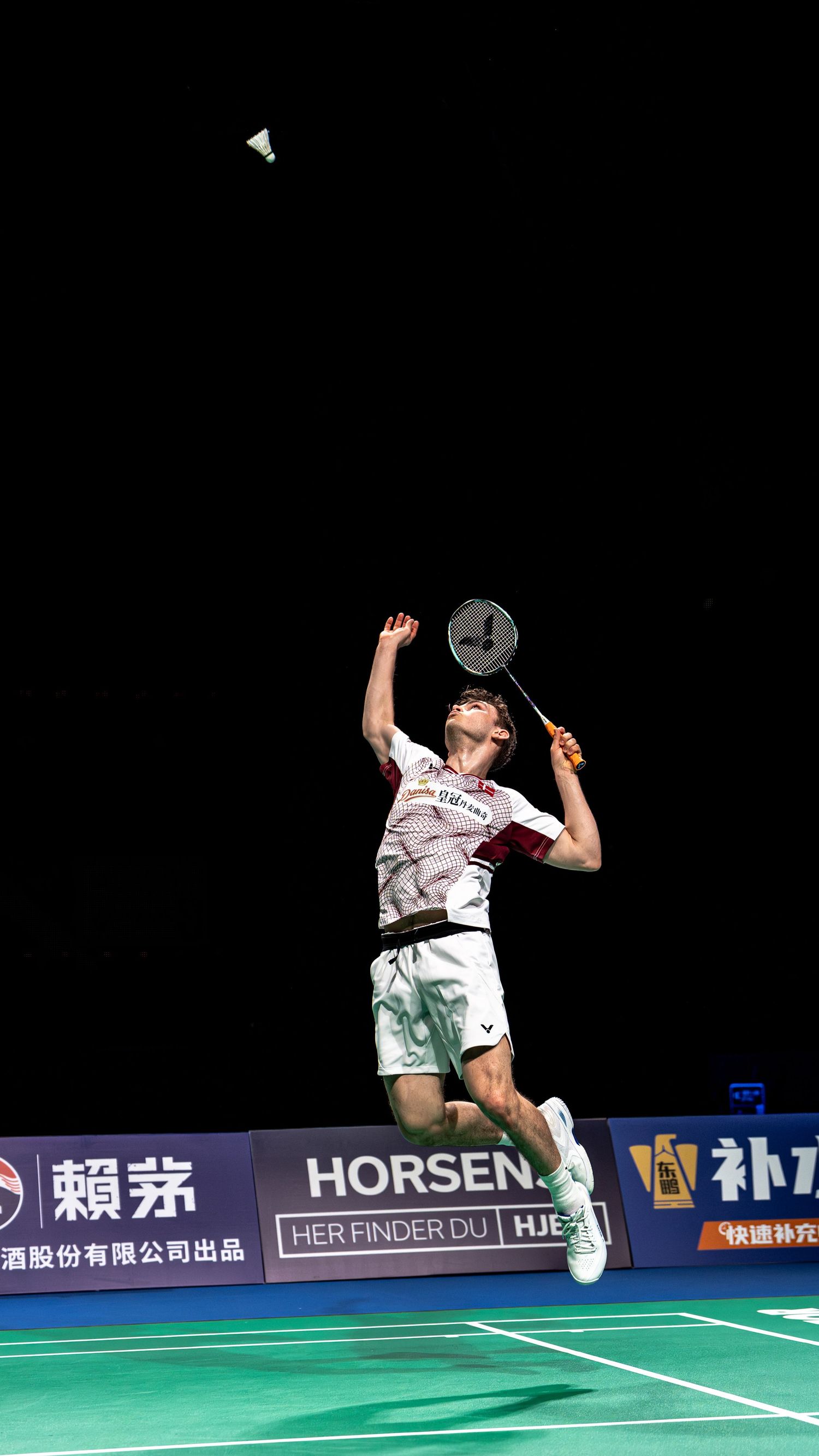 Badminton player leaping high to smash shuttlecock at indoor tournament with Horsen sponsorship banners.