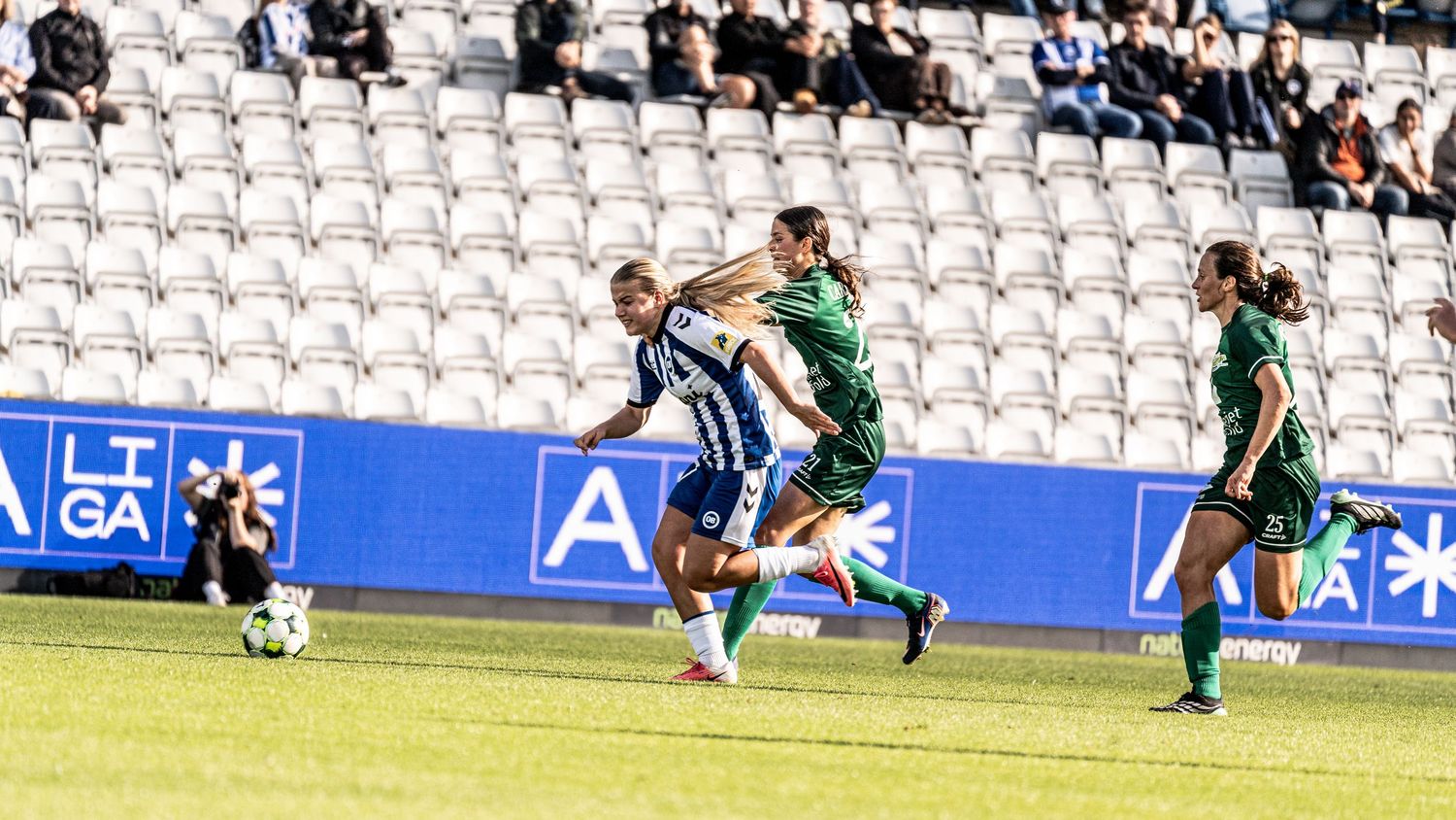 Female soccer players in blue white and green kits compete during match in stadium with sparse crowd.