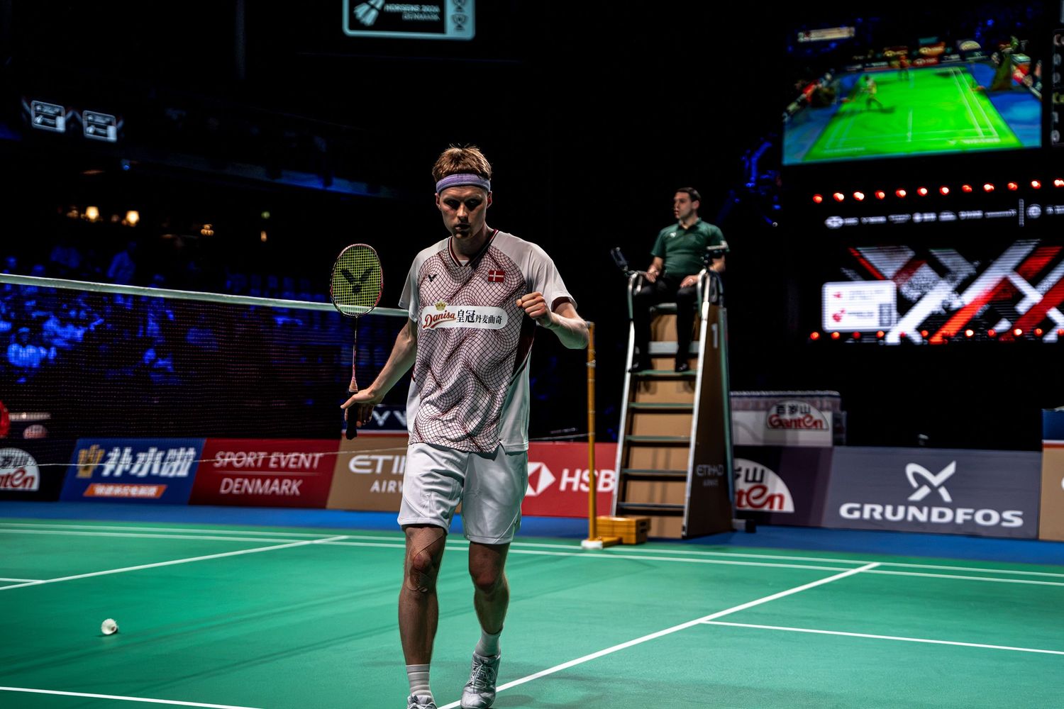 Professional badminton player mid-match on indoor court with HSBC and Grundfos sponsorship banners visible.