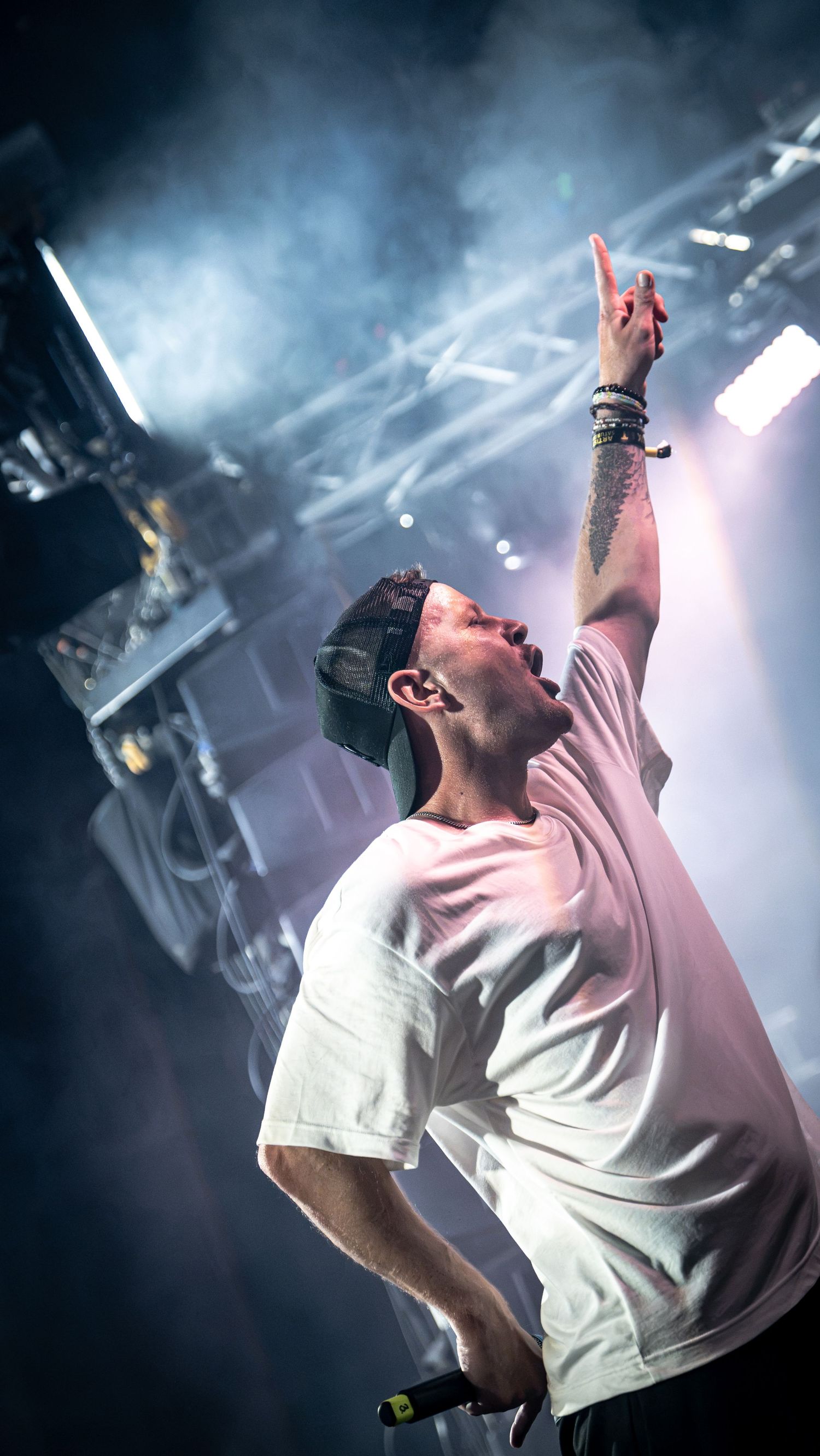 Male performer in white tee raises hand with rock gesture on smoky concert stage.