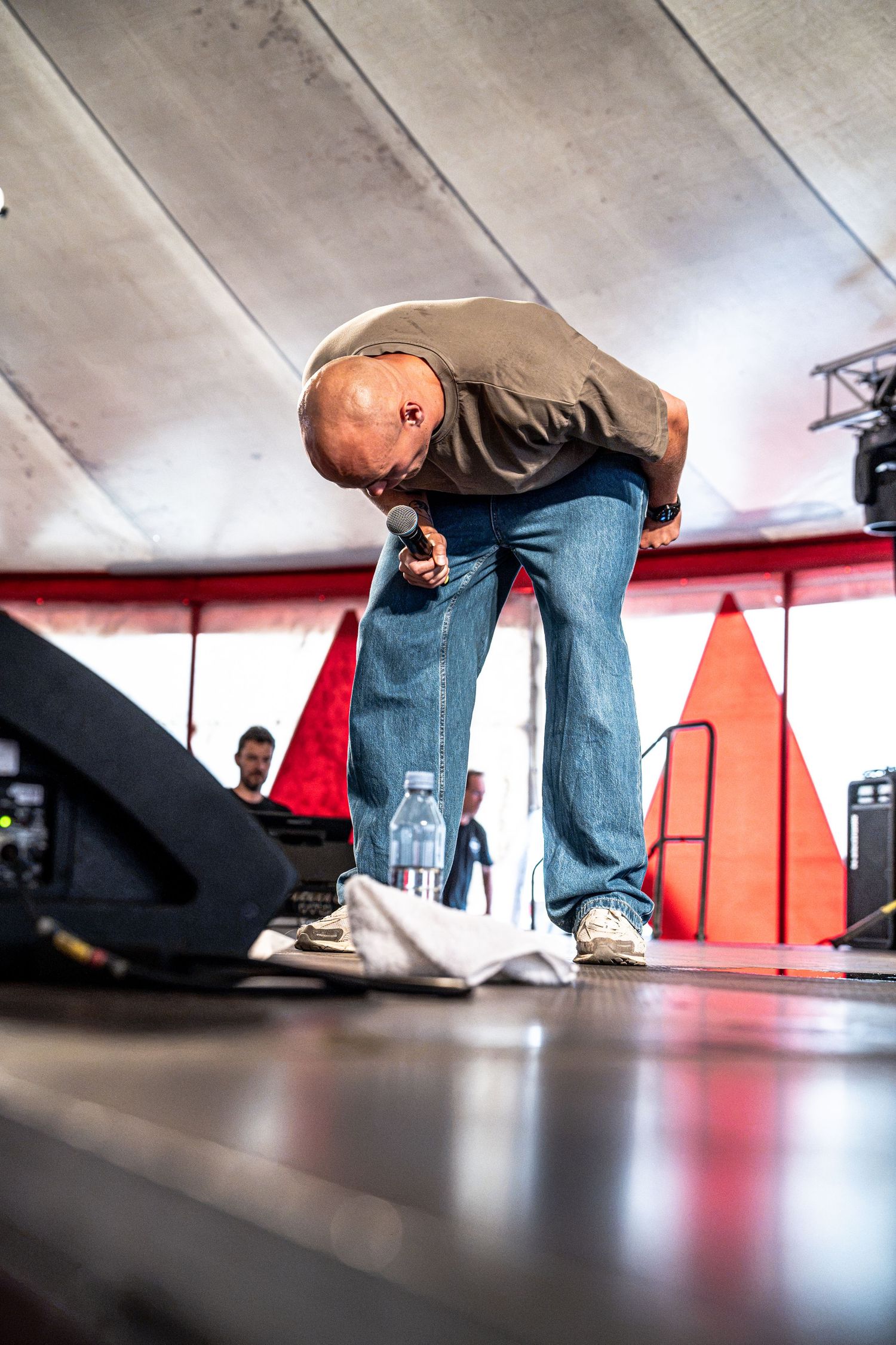 Bald performer bending forward on stage holding a microphone during a live event.