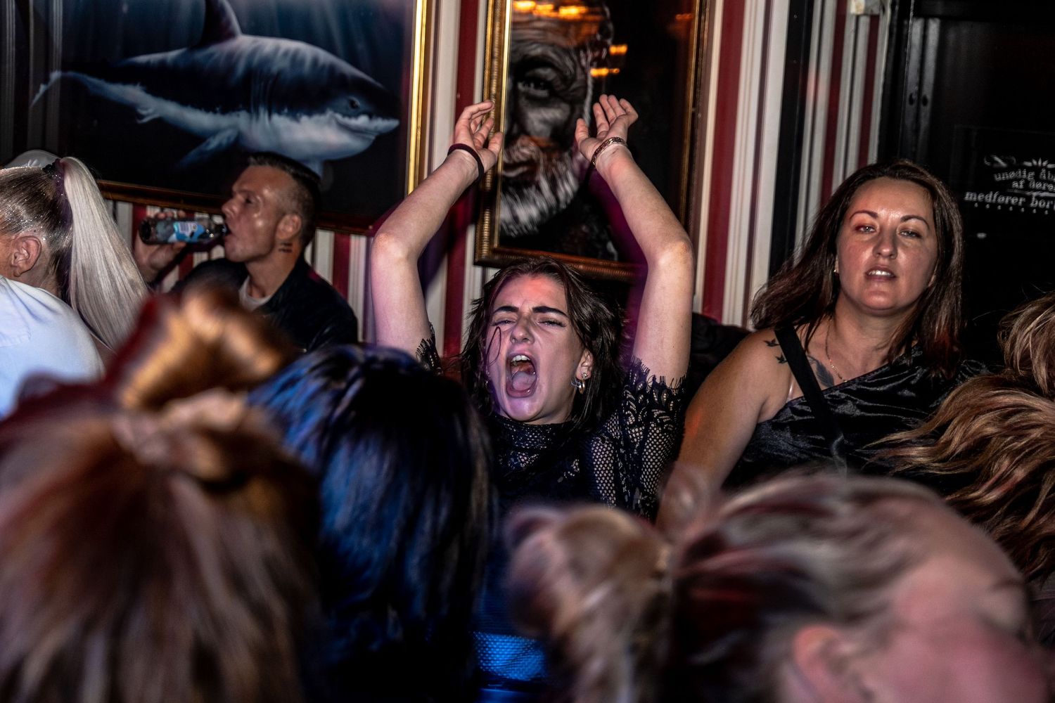 A woman with her arms raised sings or cheers loudly at a lively bar event while surrounded by a packed crowd of partygoers.