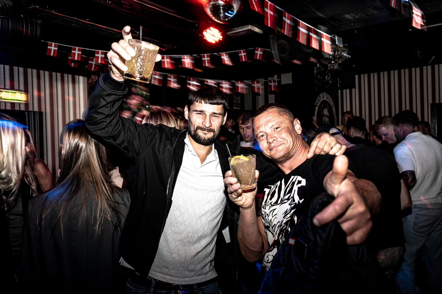 Two cheerful men raise drinks and point at the camera while celebrating in a crowded, dimly lit bar with festive bunting overhead.