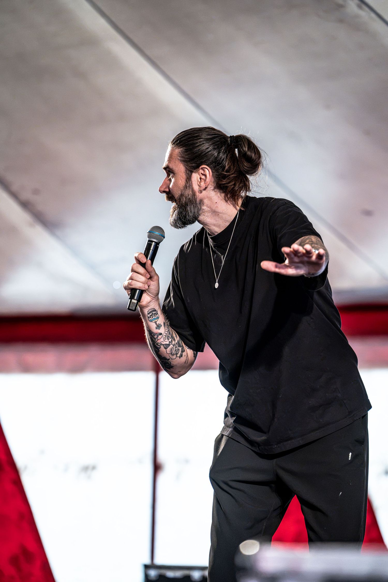 Heavily tattooed male speaker in black outfit reaching out hand while holding mic on stage.