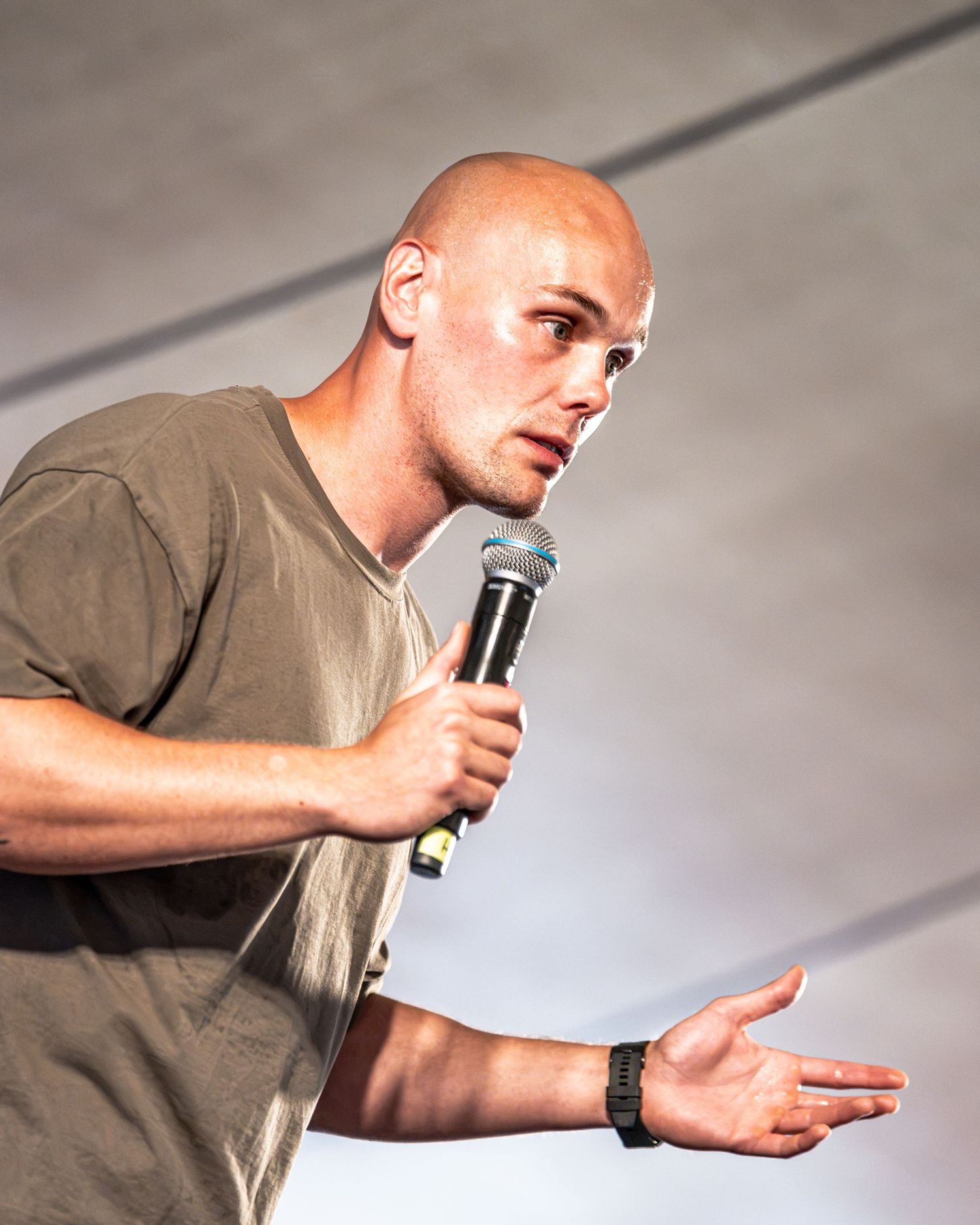 Close-up of bald man passionately speaking into microphone wearing olive t-shirt and black watch.