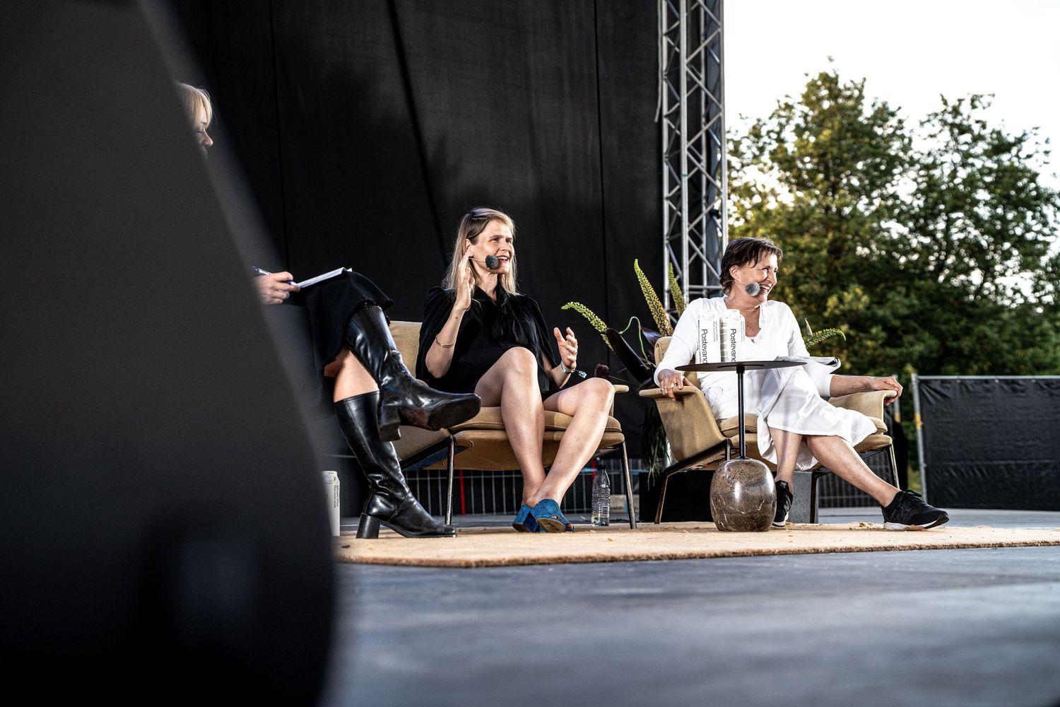 Two women seated on an outdoor stage, one in black dress touching face, one in white reading papers.