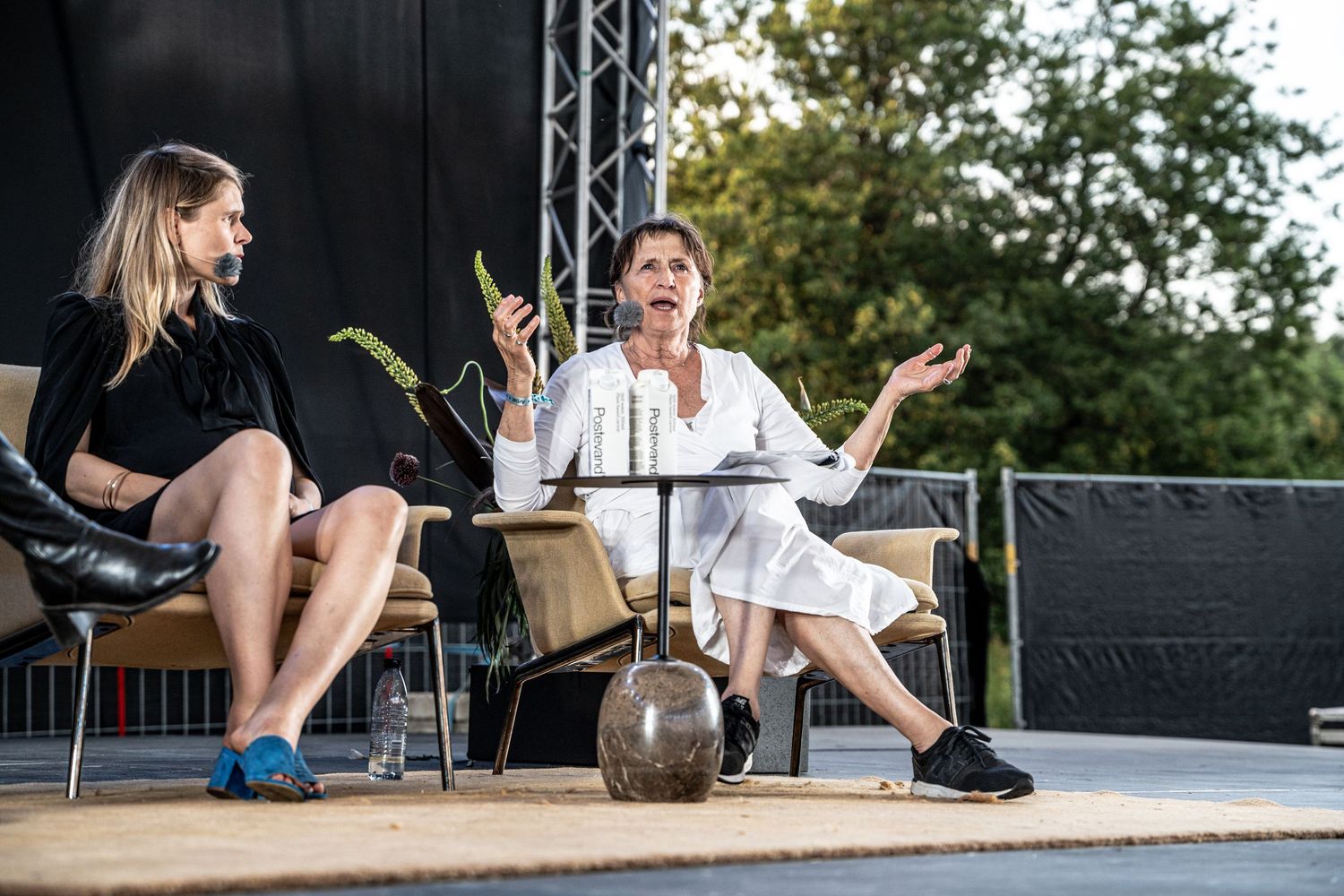 Two women in conversation on an outdoor stage, one in white gesturing with both hands.