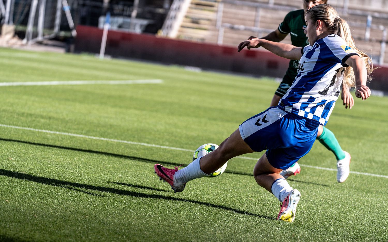 Female soccer player in blue and white kit executing a powerful kick during a competitive match on artificial turf.