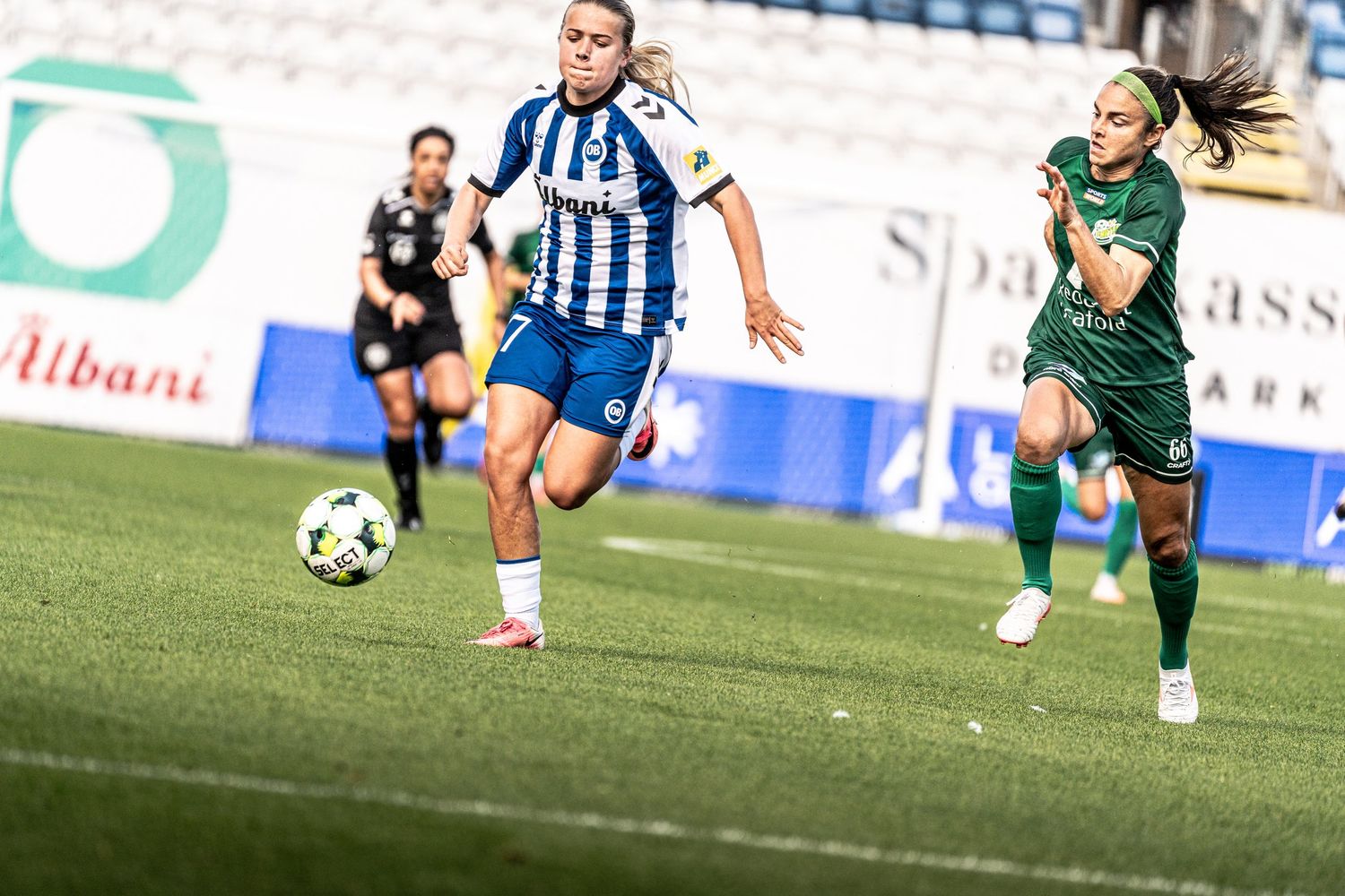 Female footballer in blue-white strip advances with ball as green-kitted opponent pursues during match.