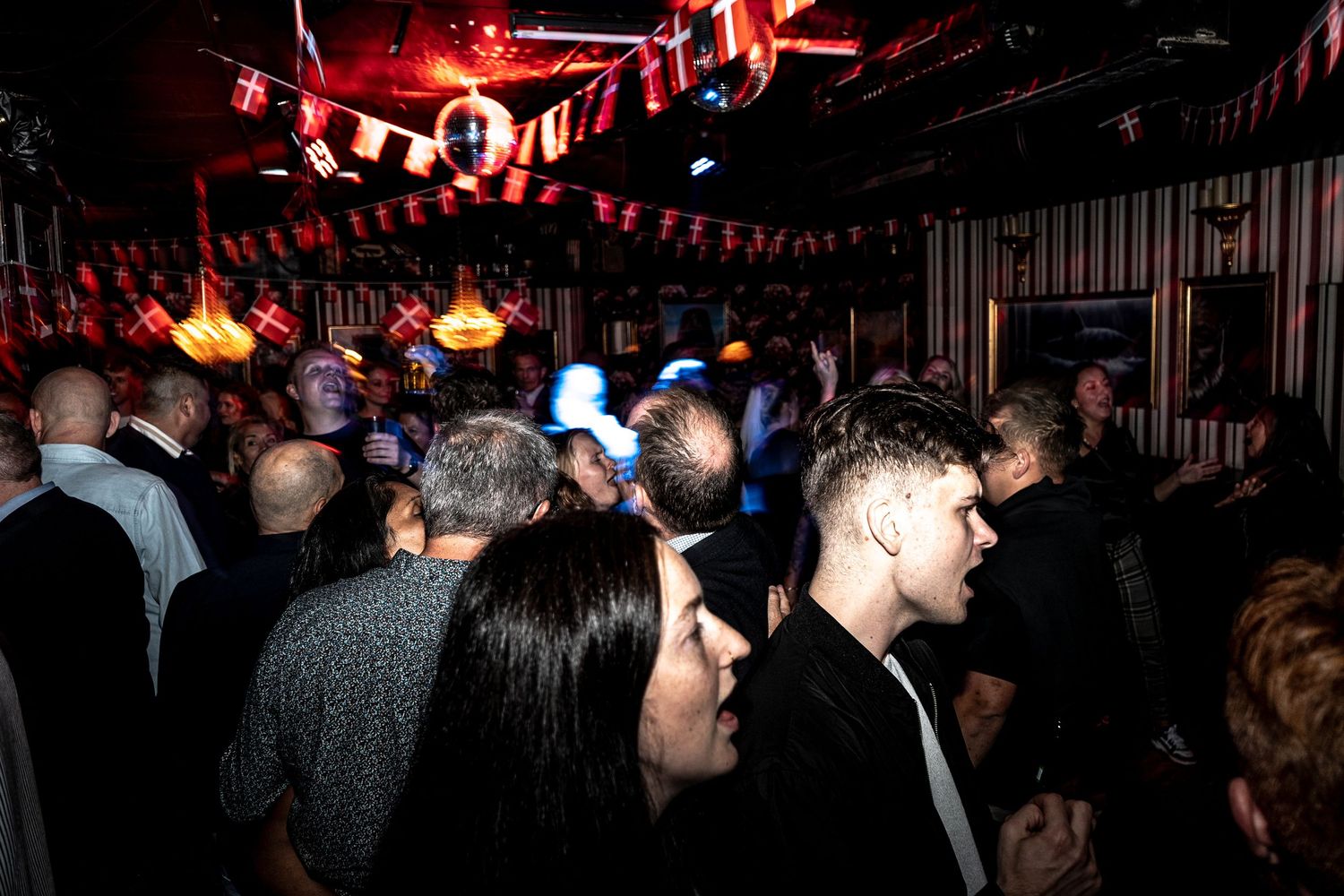 Crowded nightclub with people dancing and socializing under red bunting, a disco ball and warm amber lights in a dimly lit venue.