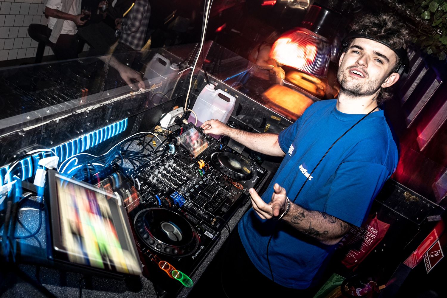 DJ in a blue t-shirt gestures while performing at a mixing console with turntables and a laptop in a dimly lit nightclub.