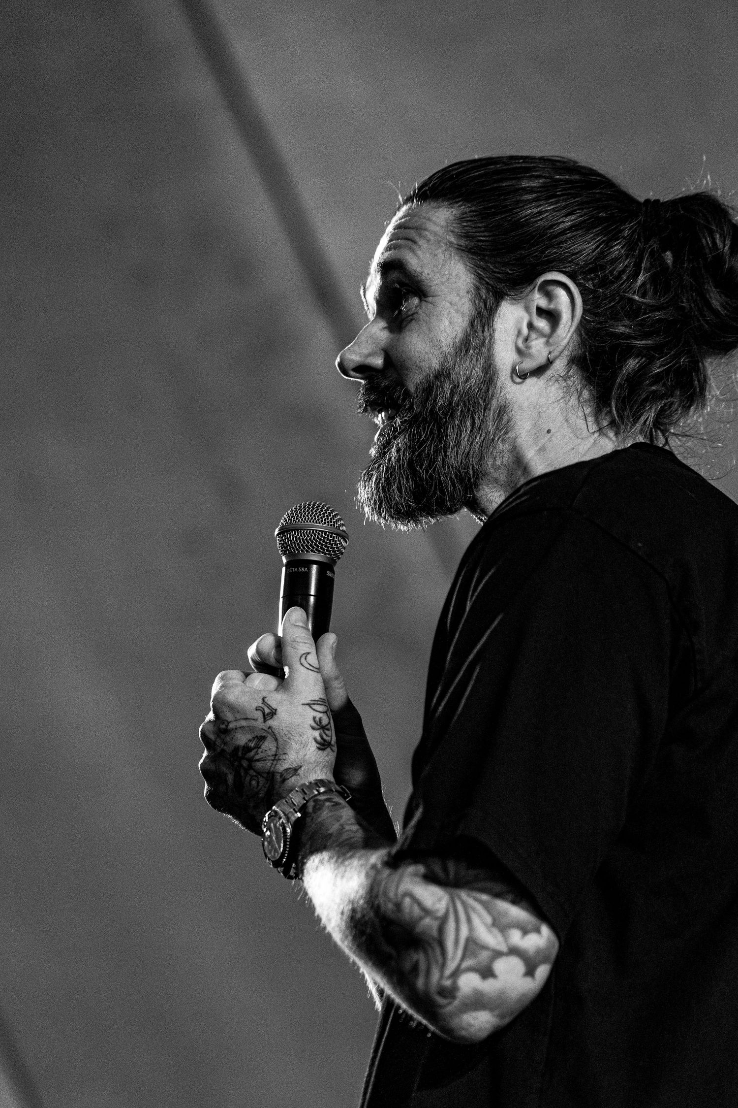 Black and white side profile of bearded tattooed man holding microphone on stage.